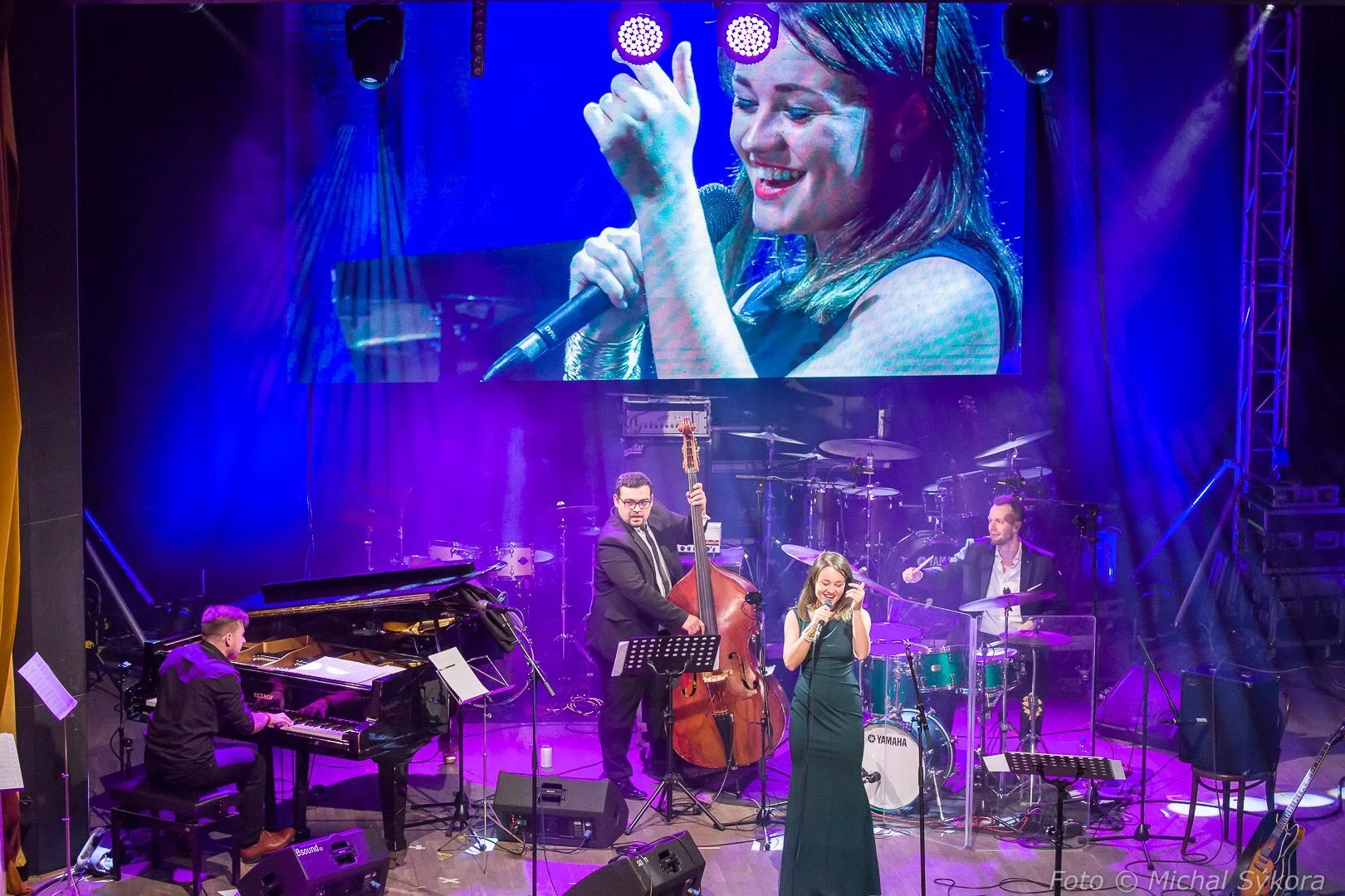 A musical performance on stage with a jazz band, including a pianist, a double bass player, a drummer, and a singer, with the singer displayed on a large screen behind them.