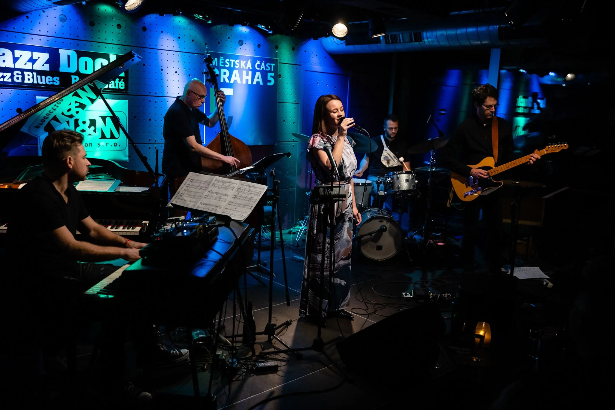 Jazz band performing live on stage with a female singer, piano, double bass, drums, and electric guitar in a dimly lit club.