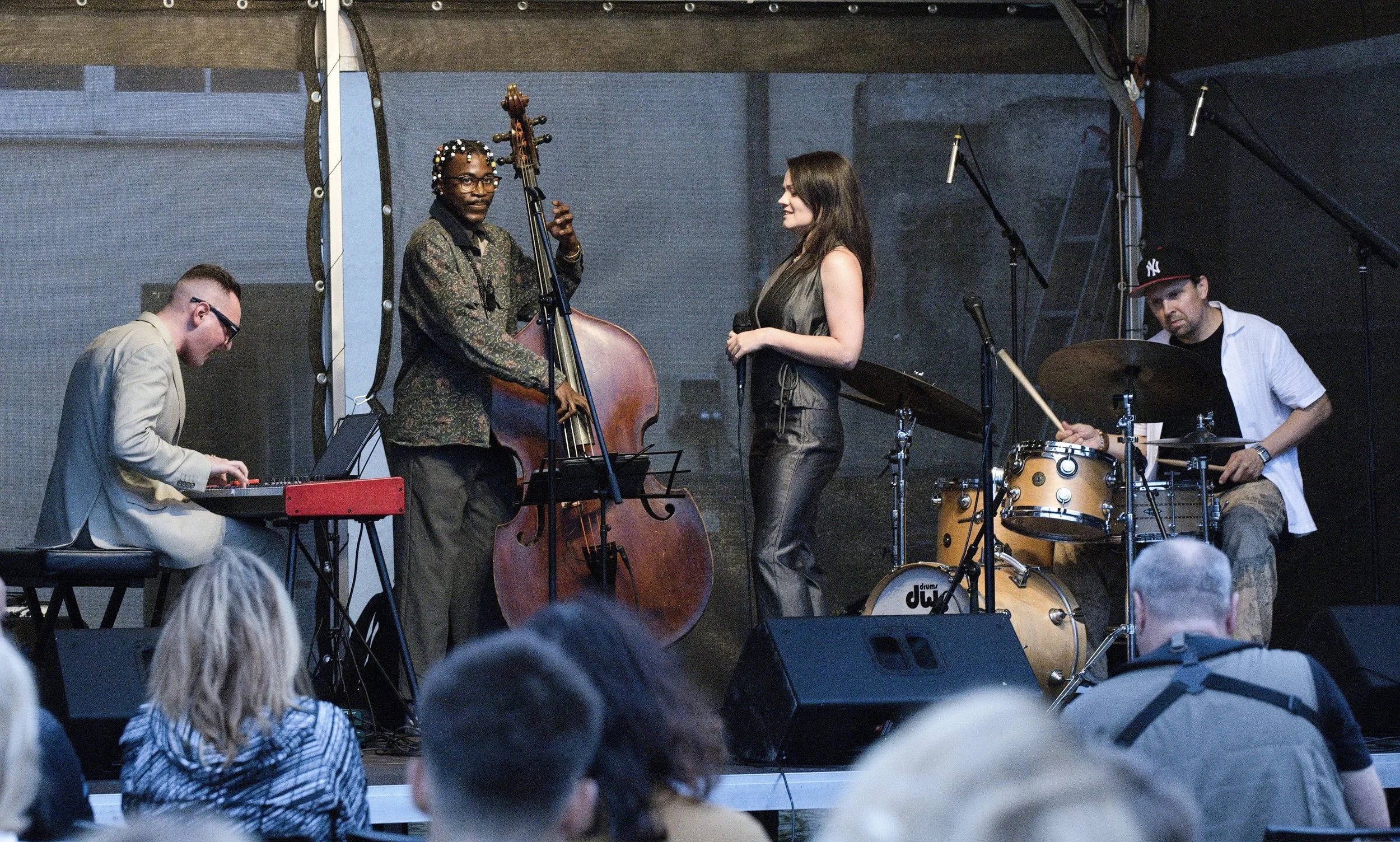 A live jazz performance on a stage with four musicians: a pianist, a double bassist, a vocalist, and a drummer. The audience is visible in the foreground.