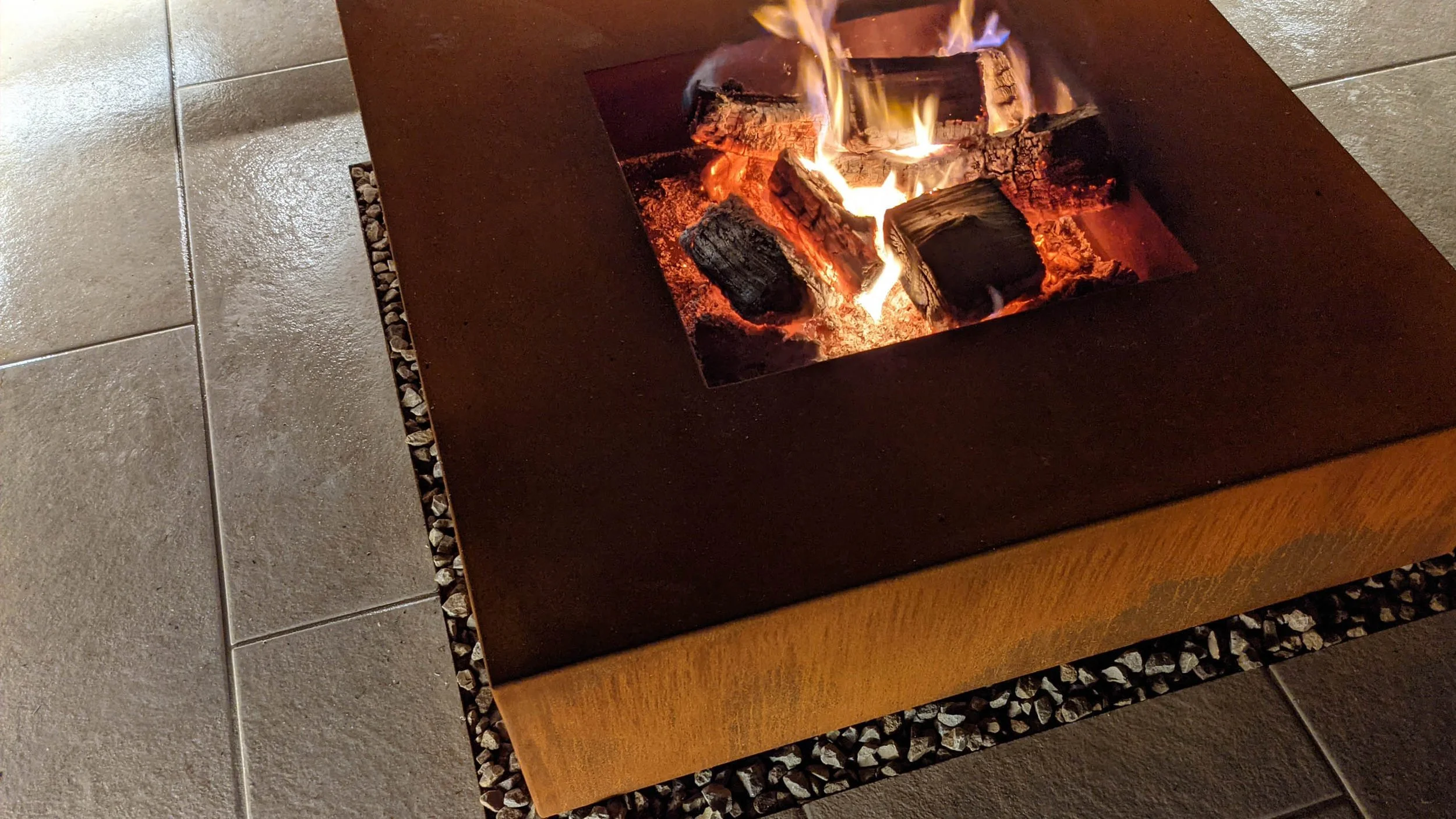 A modern tabletop fireplace built into a dark brown box with a rectangle cutout revealing burning logs and flames inside. The fireplace is set on a wooden surface with a border of small white pebbles, and the floor is made of large beige tiles.
