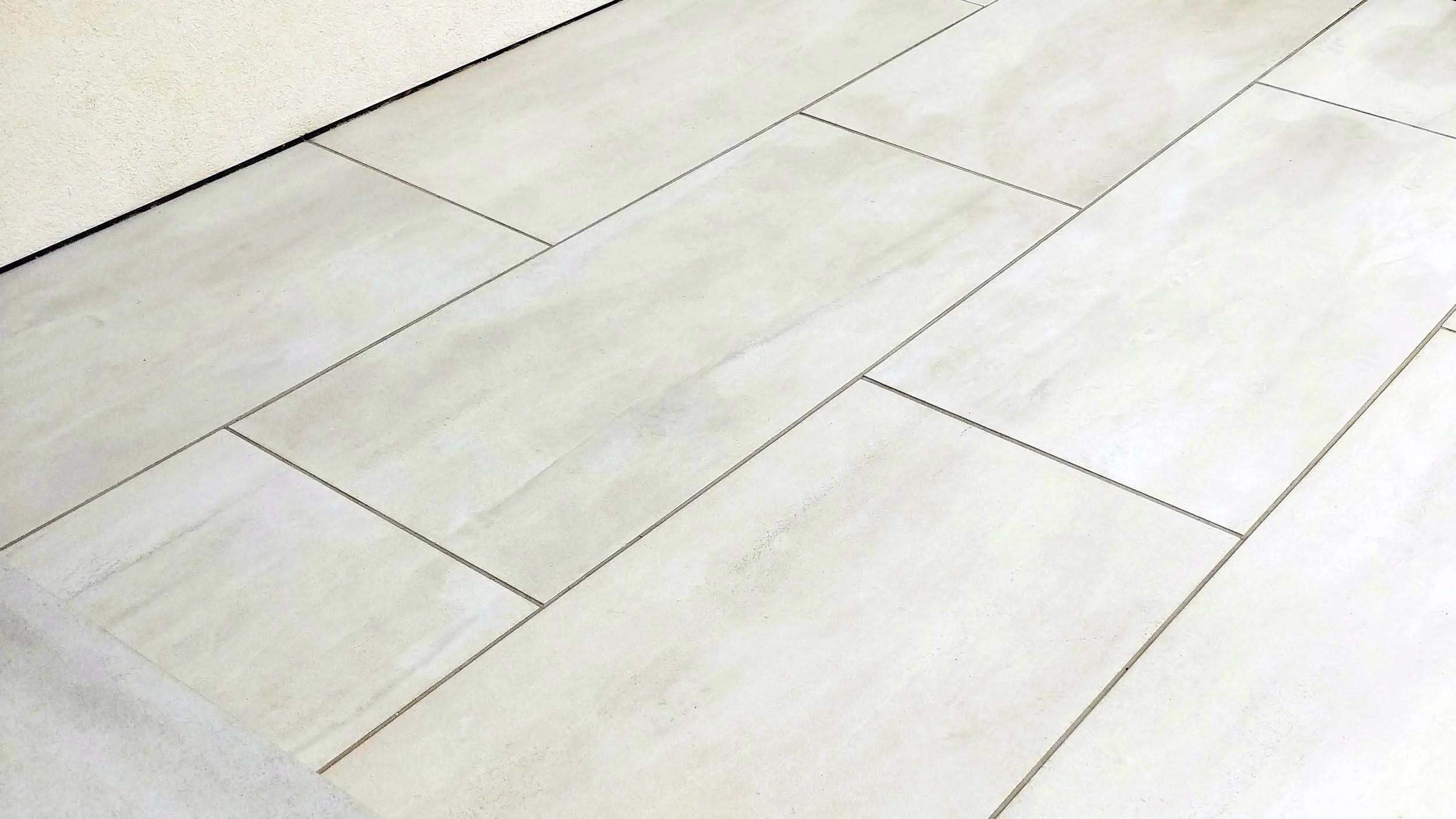 White tiled floor with light gray grout lines, partially visible beige wall with black trim along the bottom edge.