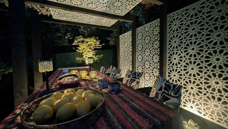 Merseyside garden dining area at night beneath a pergola, with illuminated decorative screens, a lit tree and lanterns on the table.