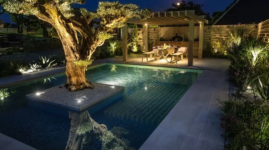 Garden pool at night with underwater lighting, a lit tree on a raised island and an illuminated dining area beneath a pergola.