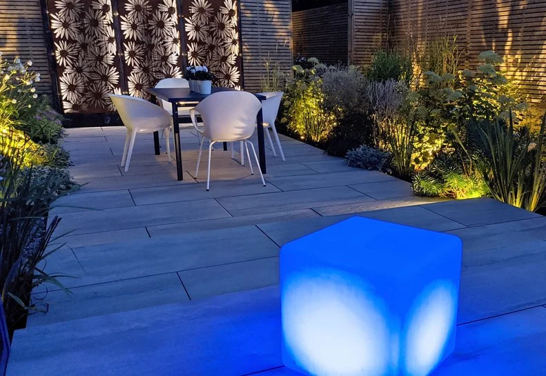 Outdoor patio with a table and six white chairs surrounded by flowering plants and illuminated by various garden lights, with a glowing blue cube in the foreground.