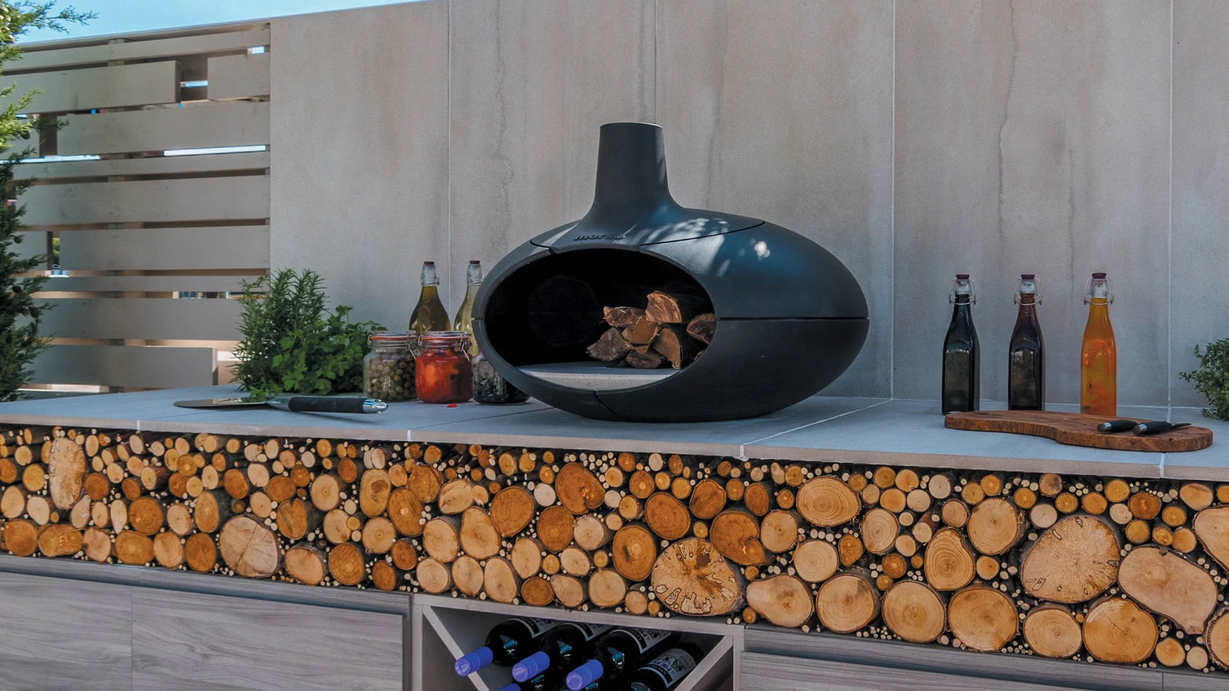 A modern outdoor kitchen with a black wood-fired oven, bottles of oil, jars of spices, fresh herbs, a cutting board with knives, and a neatly stacked firewood arrangement on a countertop.