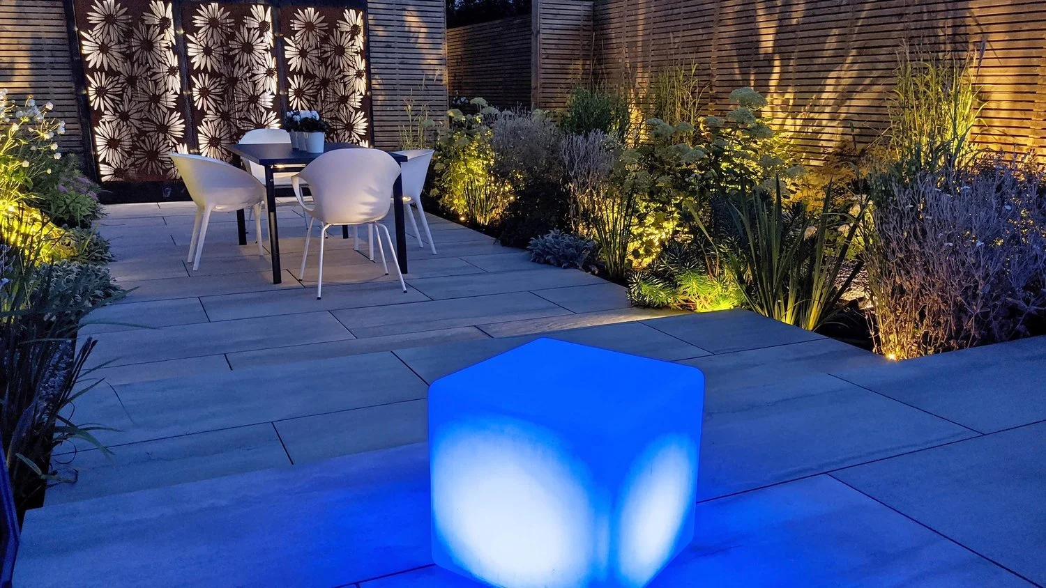 A modern outdoor patio at night with a dining table and chairs, decorative garden plants illuminated by landscape lighting, and a glowing blue cube in the foreground.