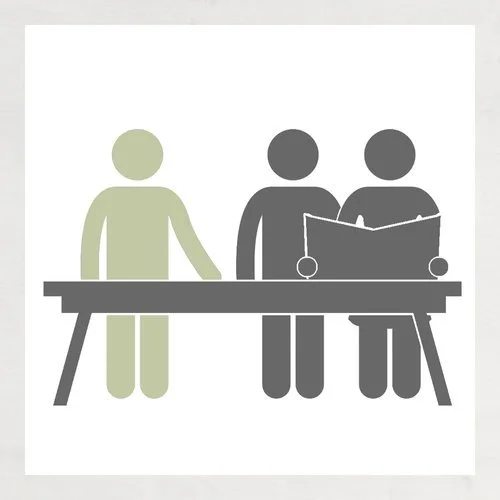 Two people standing at a table, one person reading a book or magazine while the other looks on.