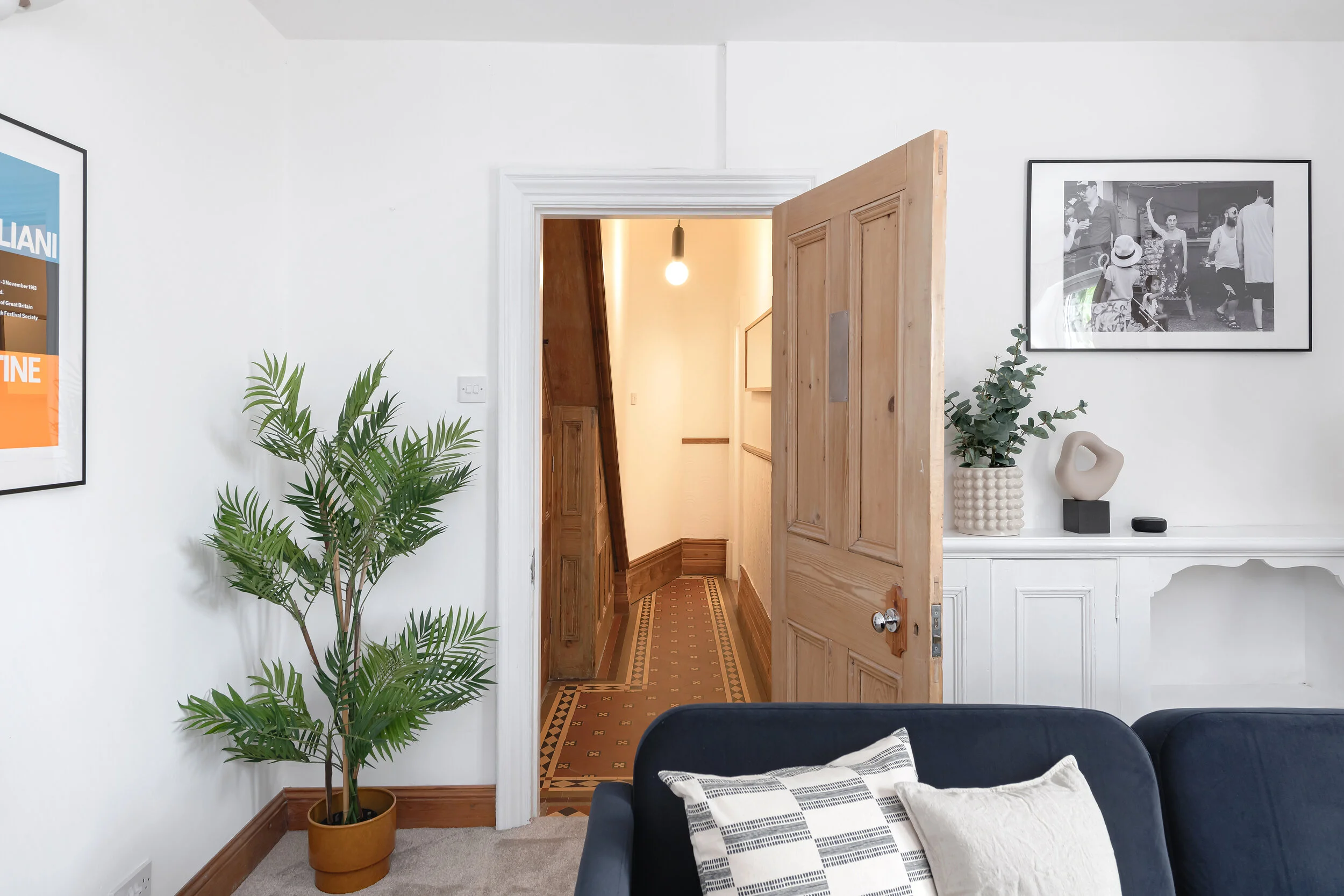 Interior view of a living room with white walls, a potted plant, a black sofa with patterned pillows, and framed black-and-white photographs on the walls. An open wooden door reveals a hallway with a patterned floor and a hanging light.
