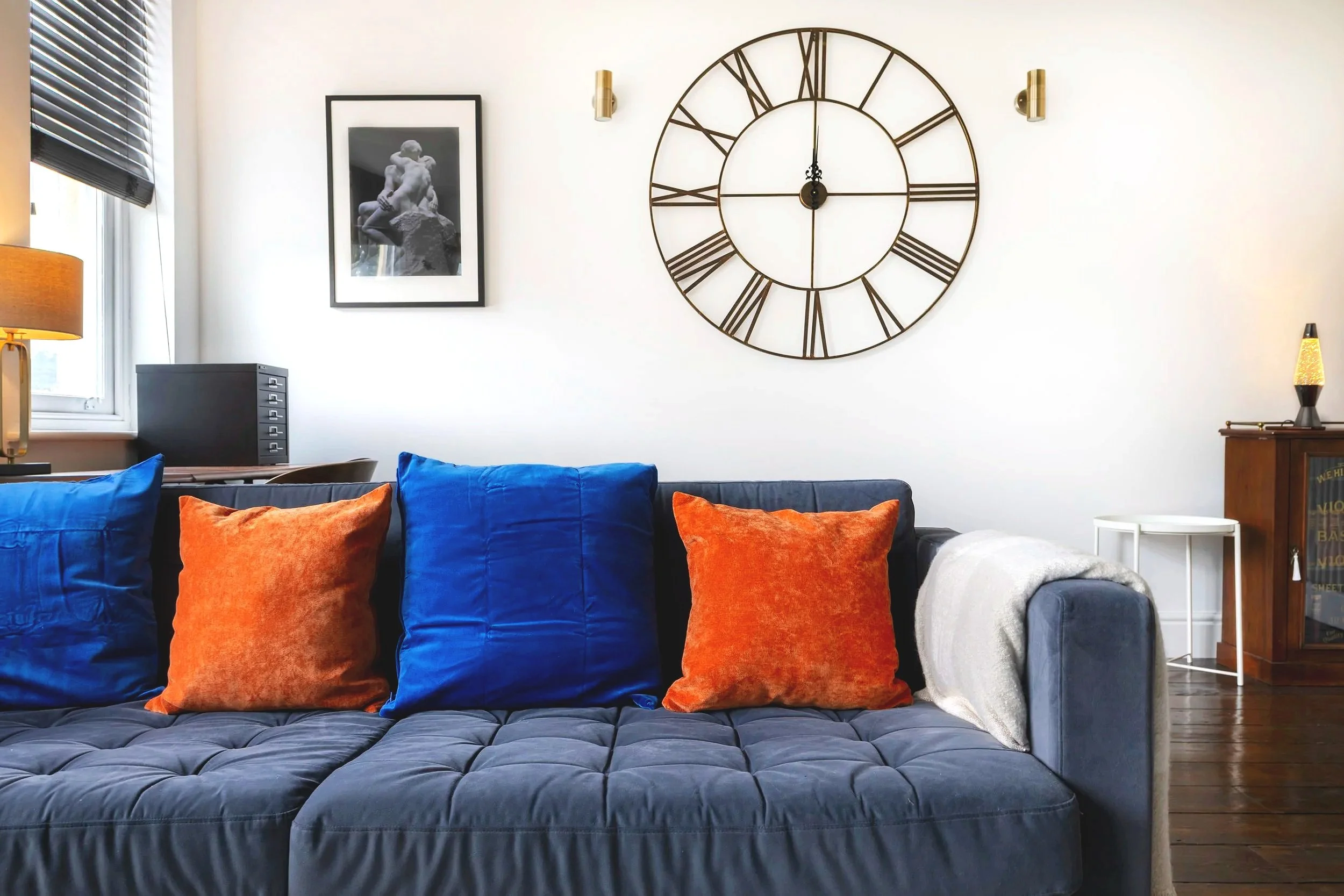 Living room with blue and orange pillows on a dark gray couch, large clock with Roman numerals on wall, framed black and white photo, window with blinds, side table and lamp, and a small cabinet with drawers.