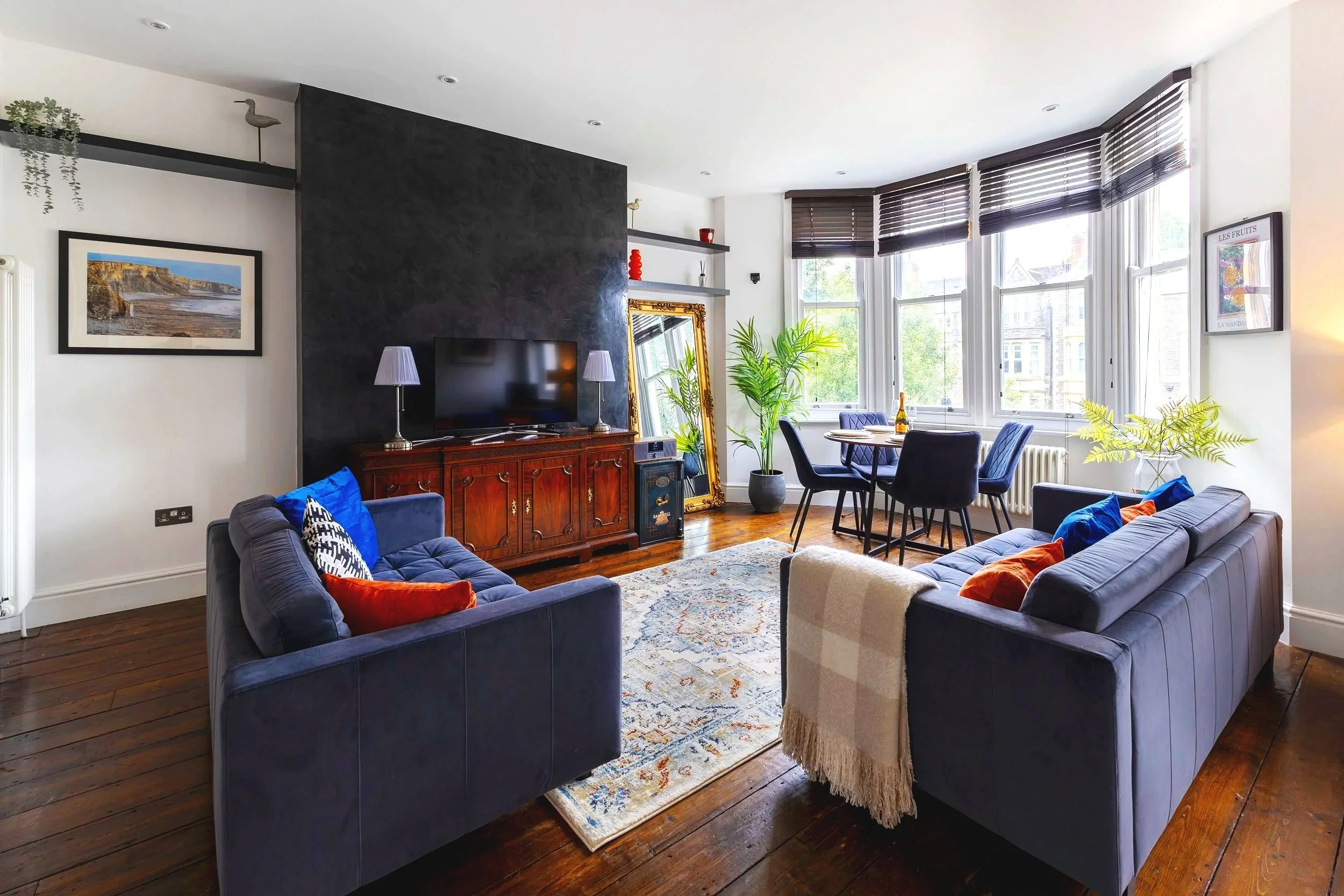 A living room with a dark blue sofa, a cream armchair with a fringed throw, a Persian-style rug, a wooden TV stand with a flat-screen TV and two purple-lamp table lamps, a large ornate gold mirror leaning against the wall, and large windows with brow