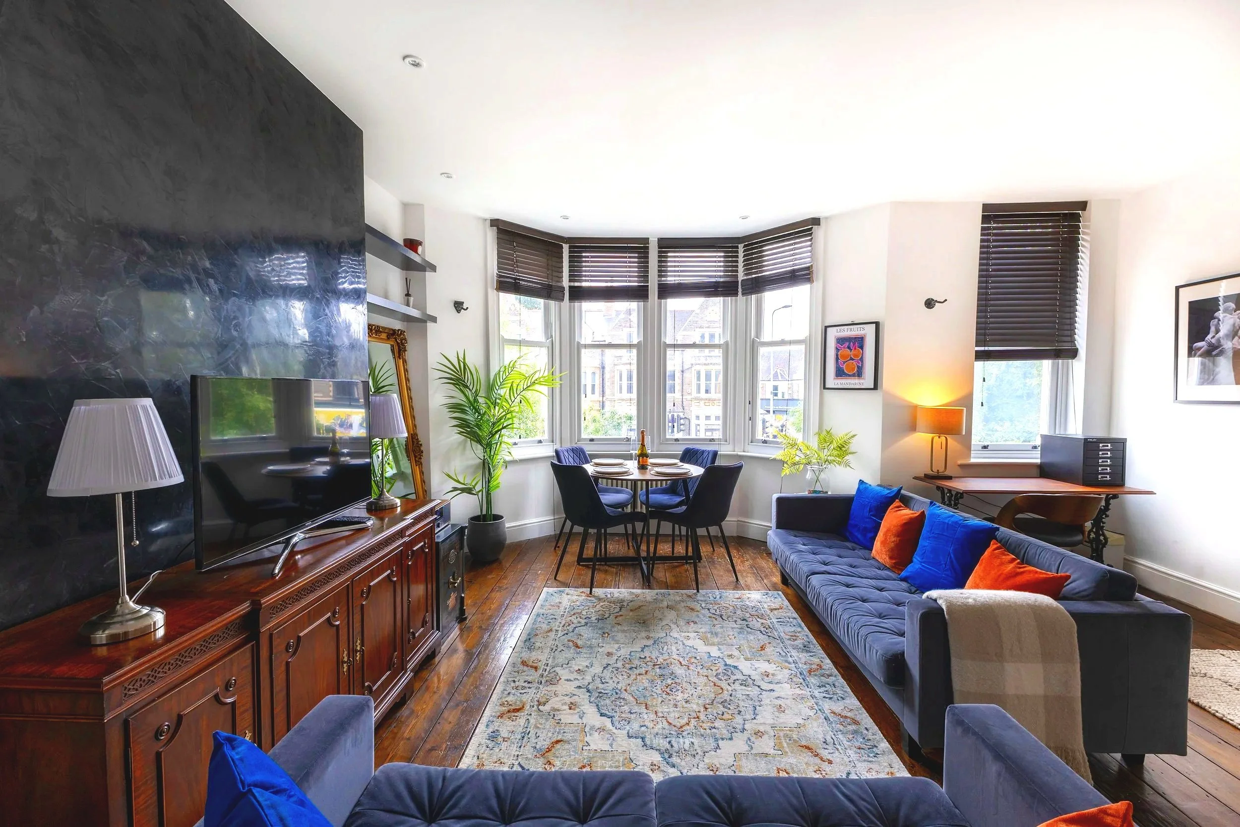 Living room with a navy blue sofa and colorful pillows, wooden coffee table, and large bay window with dark blinds. There are plants, framed pictures, and a flat-screen TV on a wooden cabinet. A round dining table with four chairs is near the windows