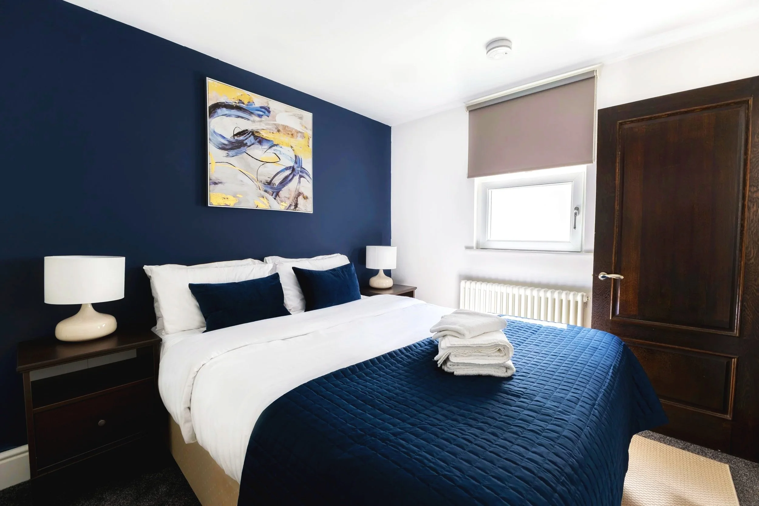 A bedroom with a dark blue accent wall and modern artwork, white bedding with navy pillows, bedside tables with white lamps, and a window with a beige roller shade, a dark wooden wardrobe, and a towel folded on the bed.