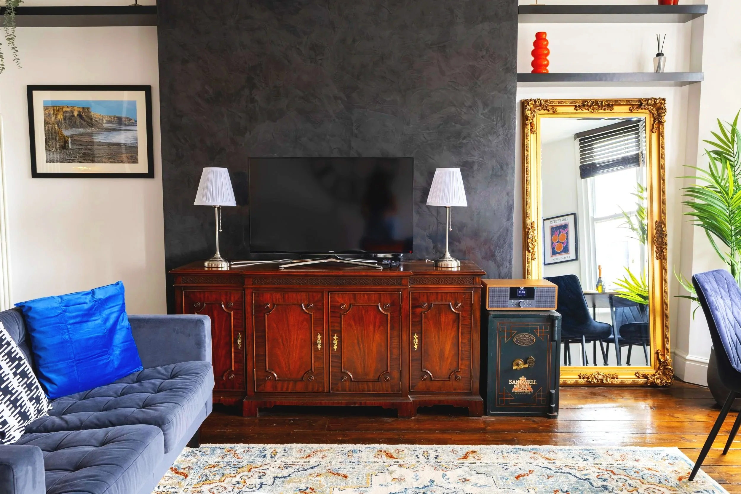 Living room with navy blue tufted sofa with blue pillows, a wooden sideboard with lamps, a large gold-framed mirror, an abstract rug, and a wall-mounted flat-screen TV. Shelves with decorative items, a window with black blinds, and a houseplant are v