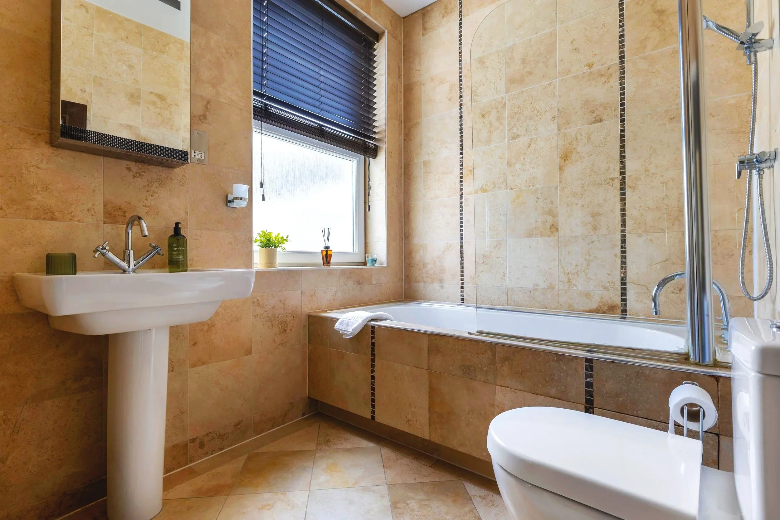 A bathroom with tan tiled walls and floor, a white sink with a chrome faucet, a mirror cabinet, a window with black blinds, and a bathtub with a glass shower door. Decor includes a small plant, a diffuser, and a soap dispenser.