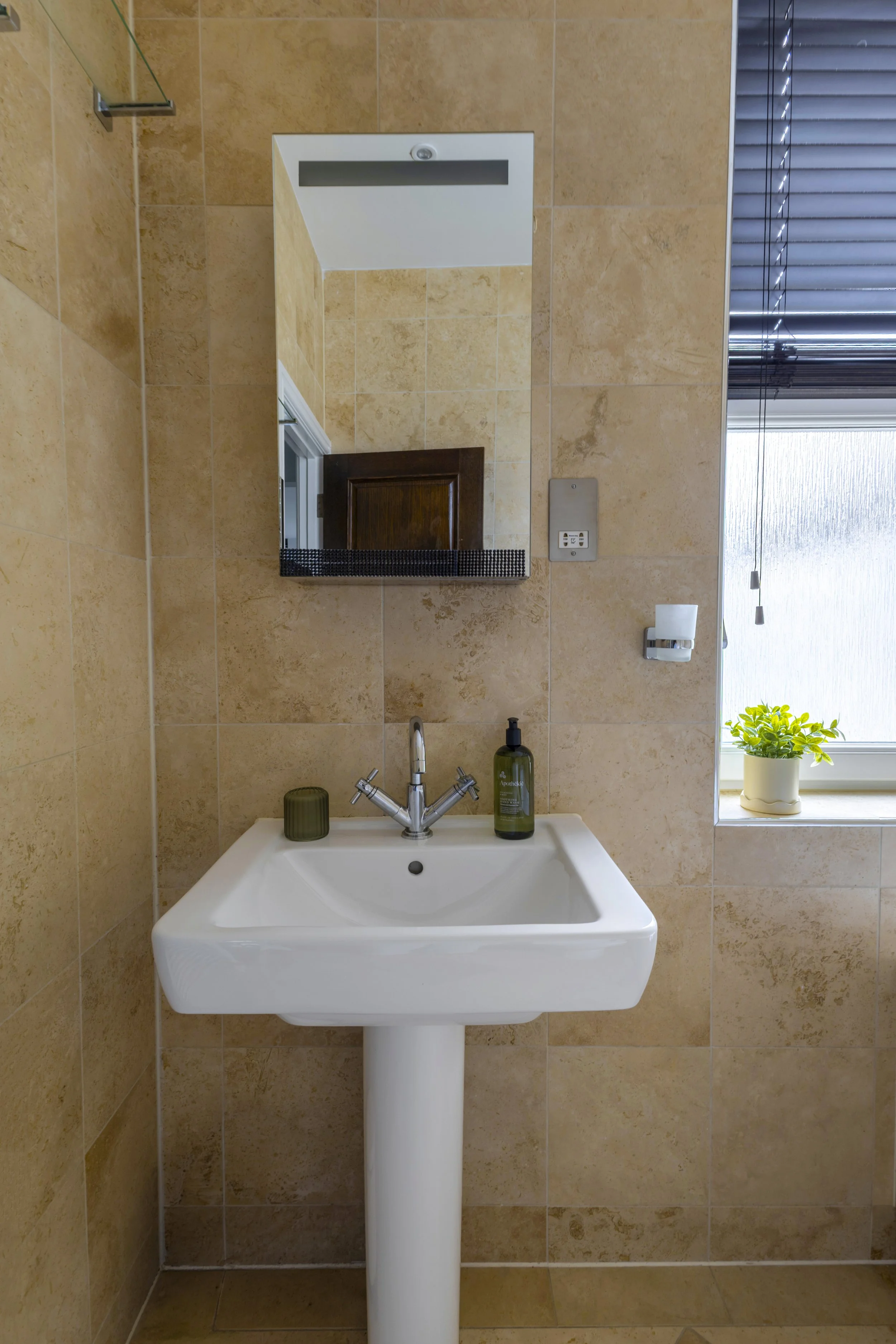 Bathroom with beige tiles, a white pedestal sink, mirror, soap dispenser, window with blinds, and a small potted plant.