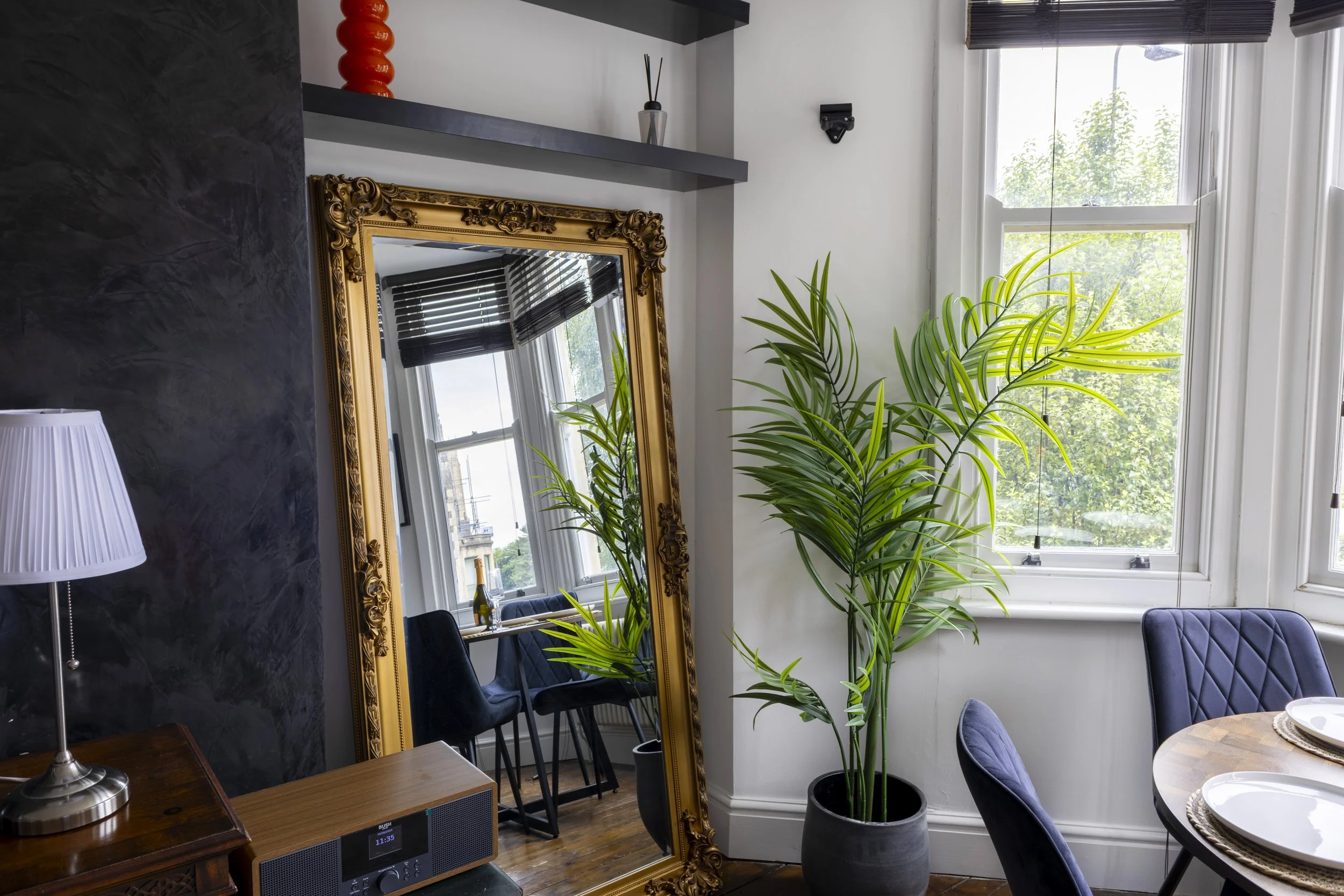 Interior of a room with a large gold-framed mirror, a potted palm tree, a window with blinds, and a small table with plates.