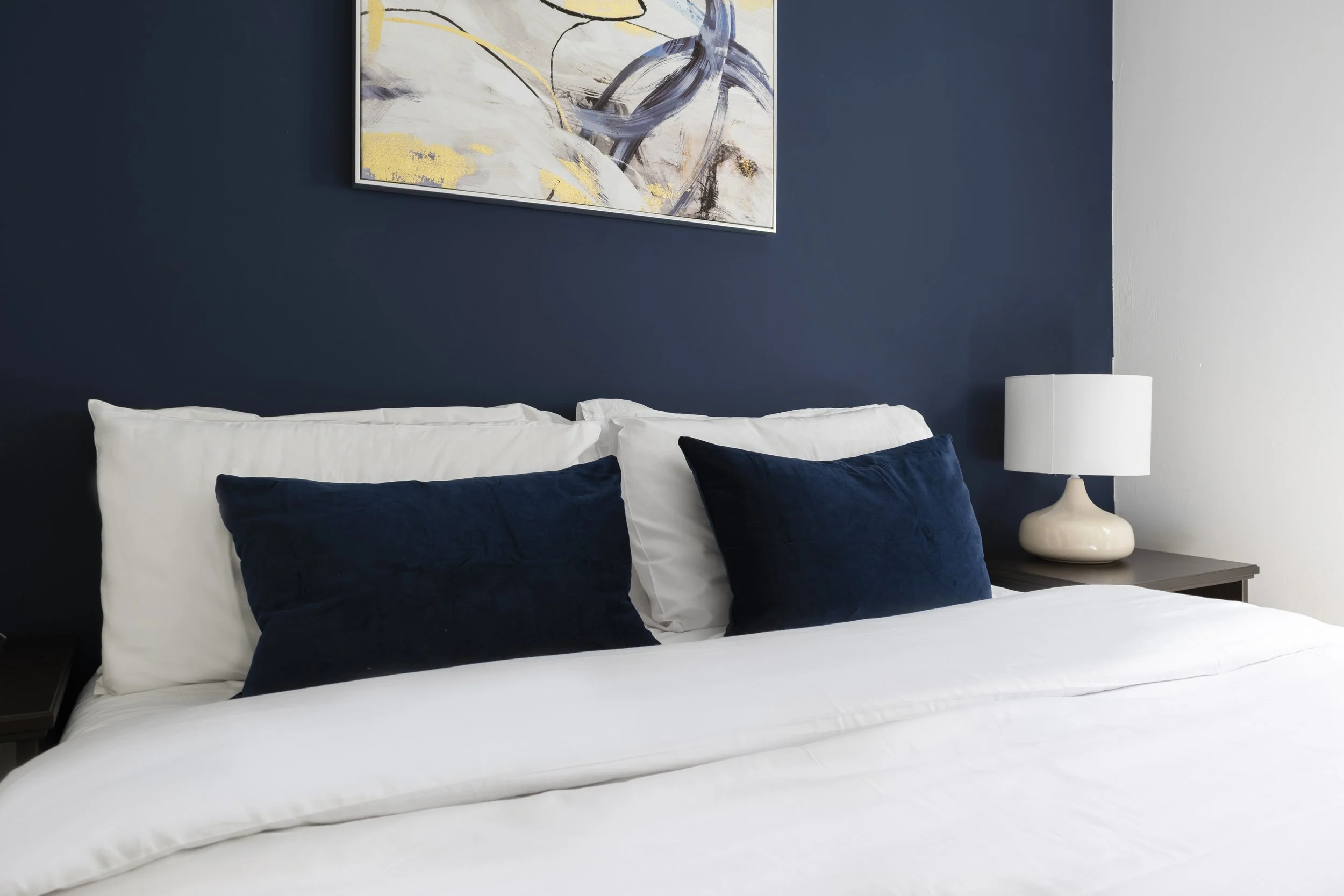 A neatly made bed with white sheets and multiple navy blue pillows, set against a dark blue accent wall. There is a white table lamp with a rounded base on a nightstand to the right of the bed and an abstract painting with shades of white, gray, blac
