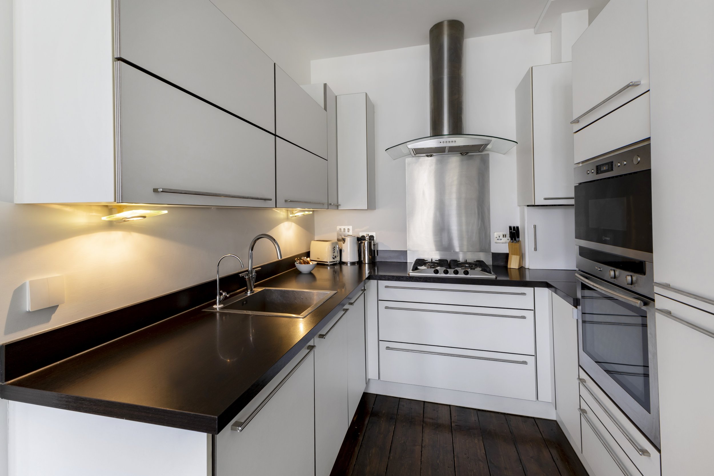 Modern kitchen with white cabinets, black countertops, and stainless steel appliances, including a stove, oven, and microwave. There is a stainless steel range hood above the stove, and under-cabinet lighting illuminates the countertop.