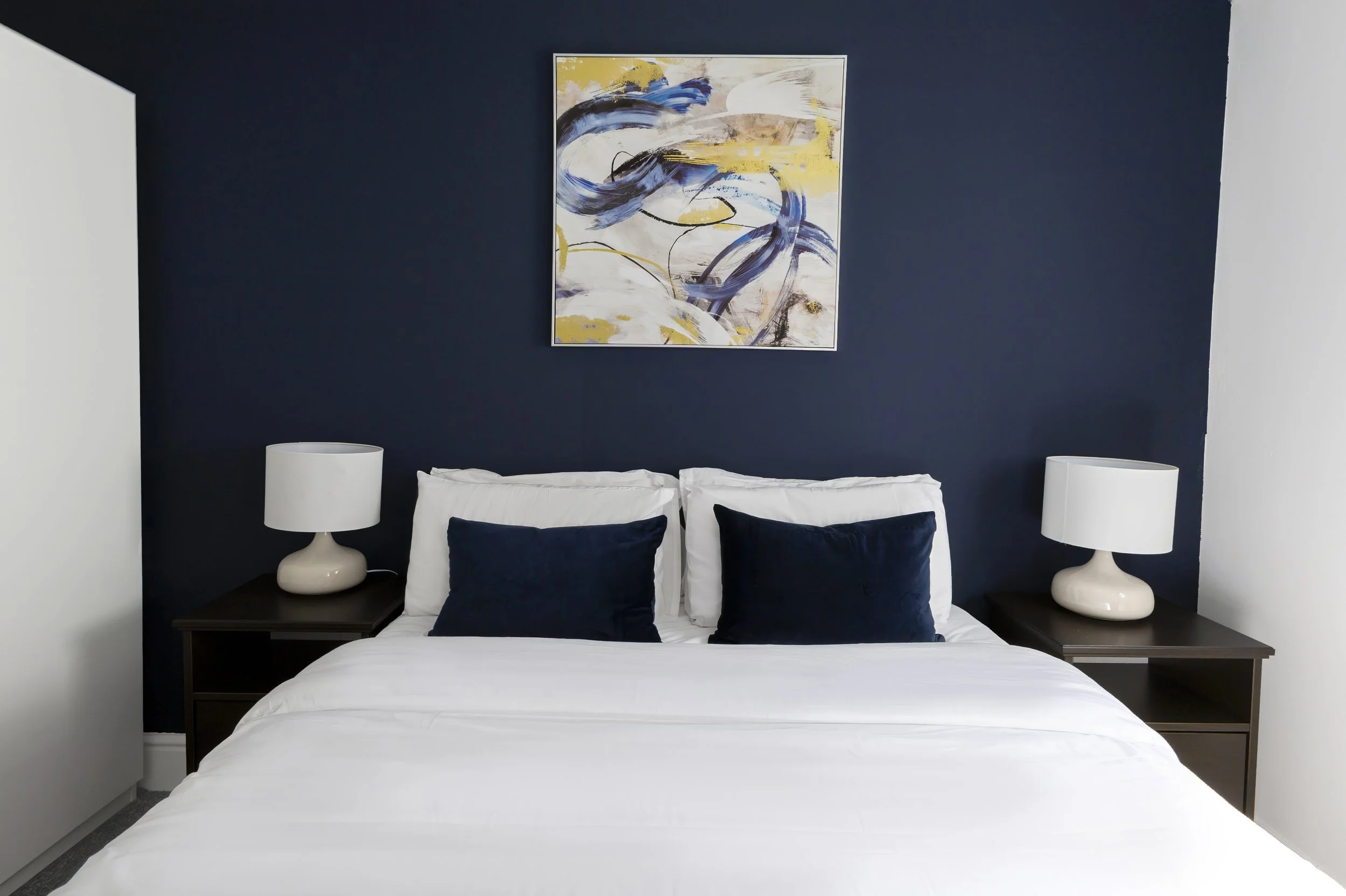 A bedroom with a bed, four pillows, two nightstands, and two white table lamps on either side of the bed. There is abstract artwork with blue, yellow, and black strokes hanging on a dark navy blue wall.