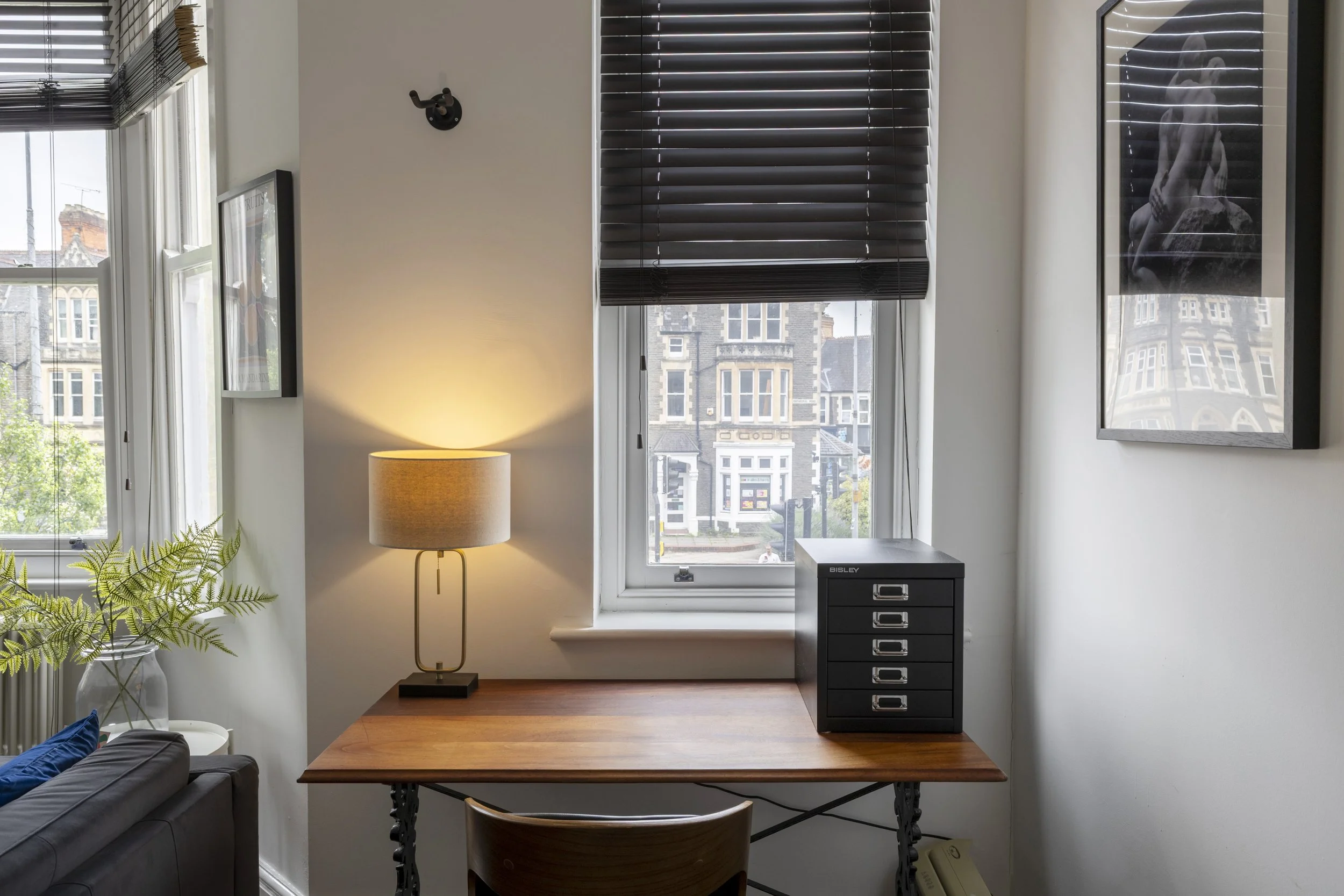 A corner of a room with a window with black blinds, a lamp with a beige shade, a black filing cabinet, a framed black and white photograph, a wooden table, a dark-colored chair, and a potted fern plant.