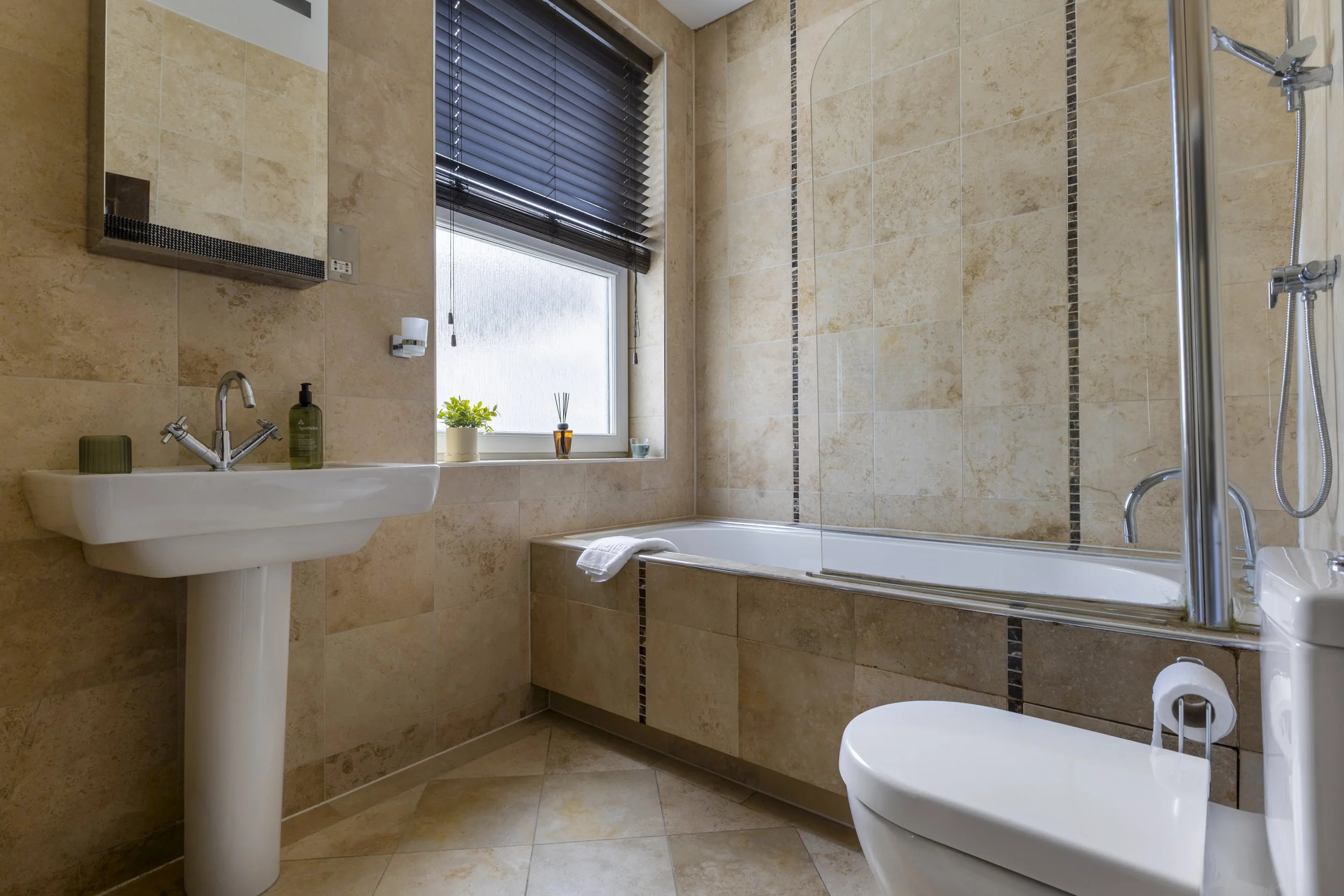A modern bathroom with beige tiled walls and floor, a white sink with a chrome faucet, a mirror cabinet, a window with black blinds, a potted plant, and a bathtub with a shower fixture.