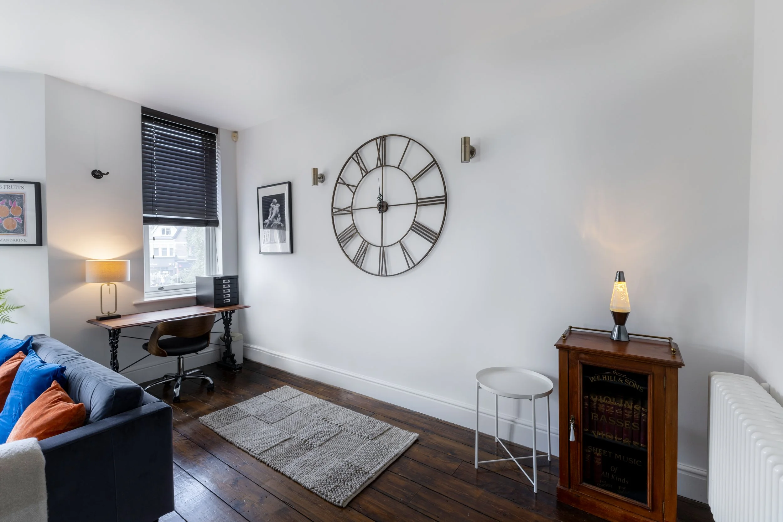 Living room with white walls, a large decorative wall clock, framed art, window with black blinds, a table with a lamp, a chair, a small white round table, and a wooden bookshelf with books and decorative items.