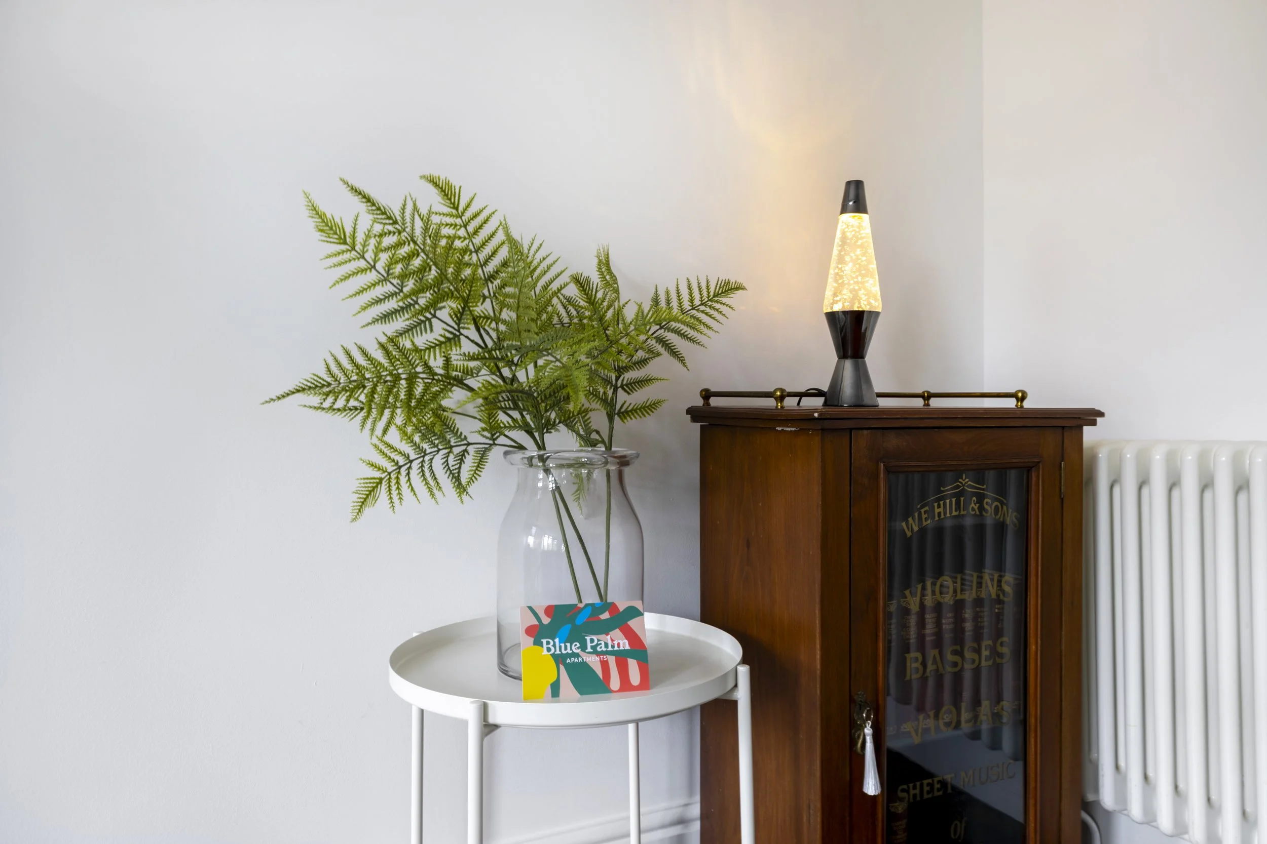 A white round table holding a clear glass vase with green fern leaves and a colorful photo card that reads 'Blue Palm Apartments'. To the right, there is a wooden cabinet with a black lava lamp on top, and a white radiator partially visible to the fa