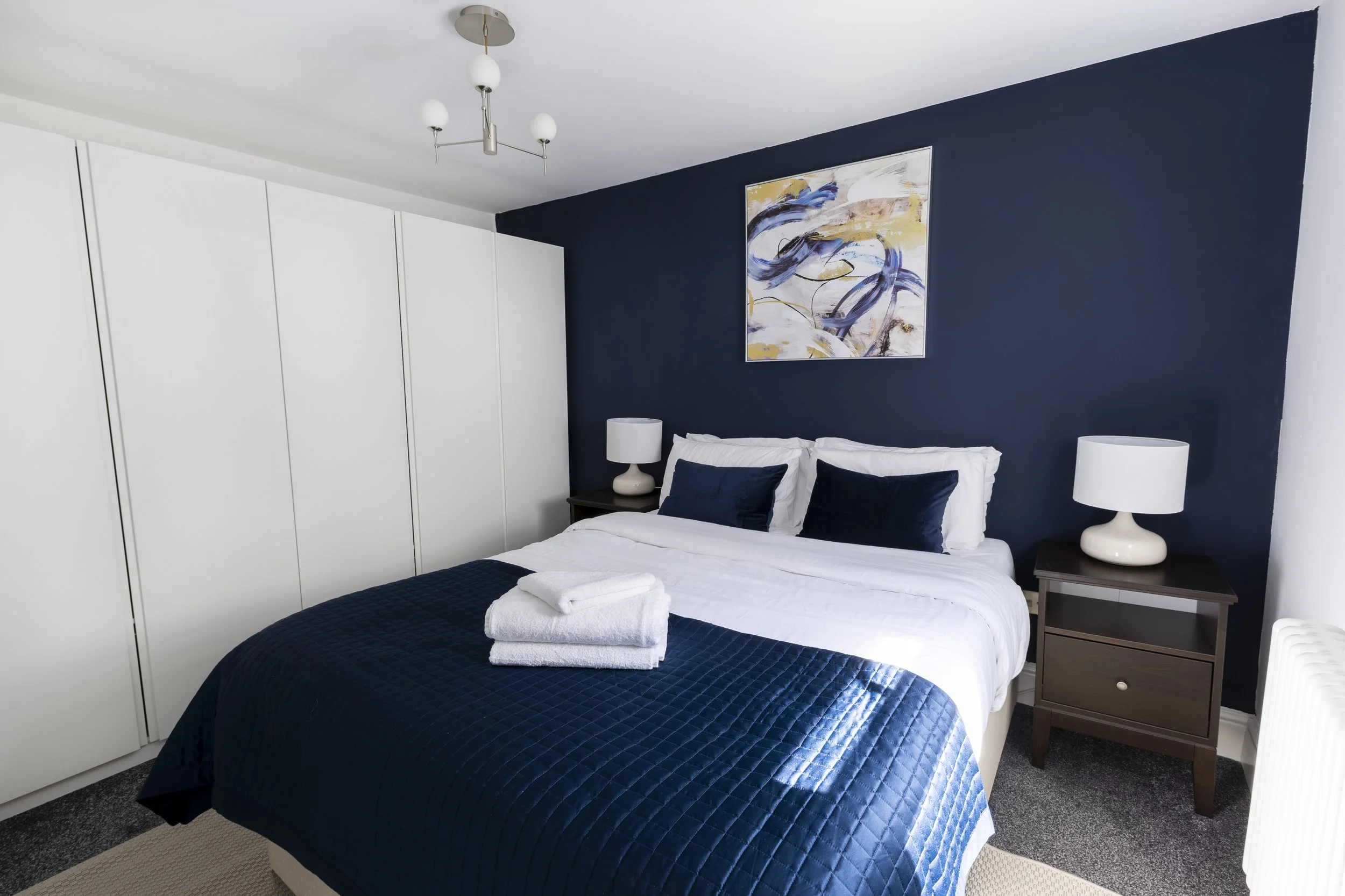 A neatly made bed with white sheets and navy blue quilt in a bedroom with navy accent wall. There are two matching navy blue pillows and two white towels on the bed. Flanking the bed are side tables with white lamps and a colorful abstract painting h