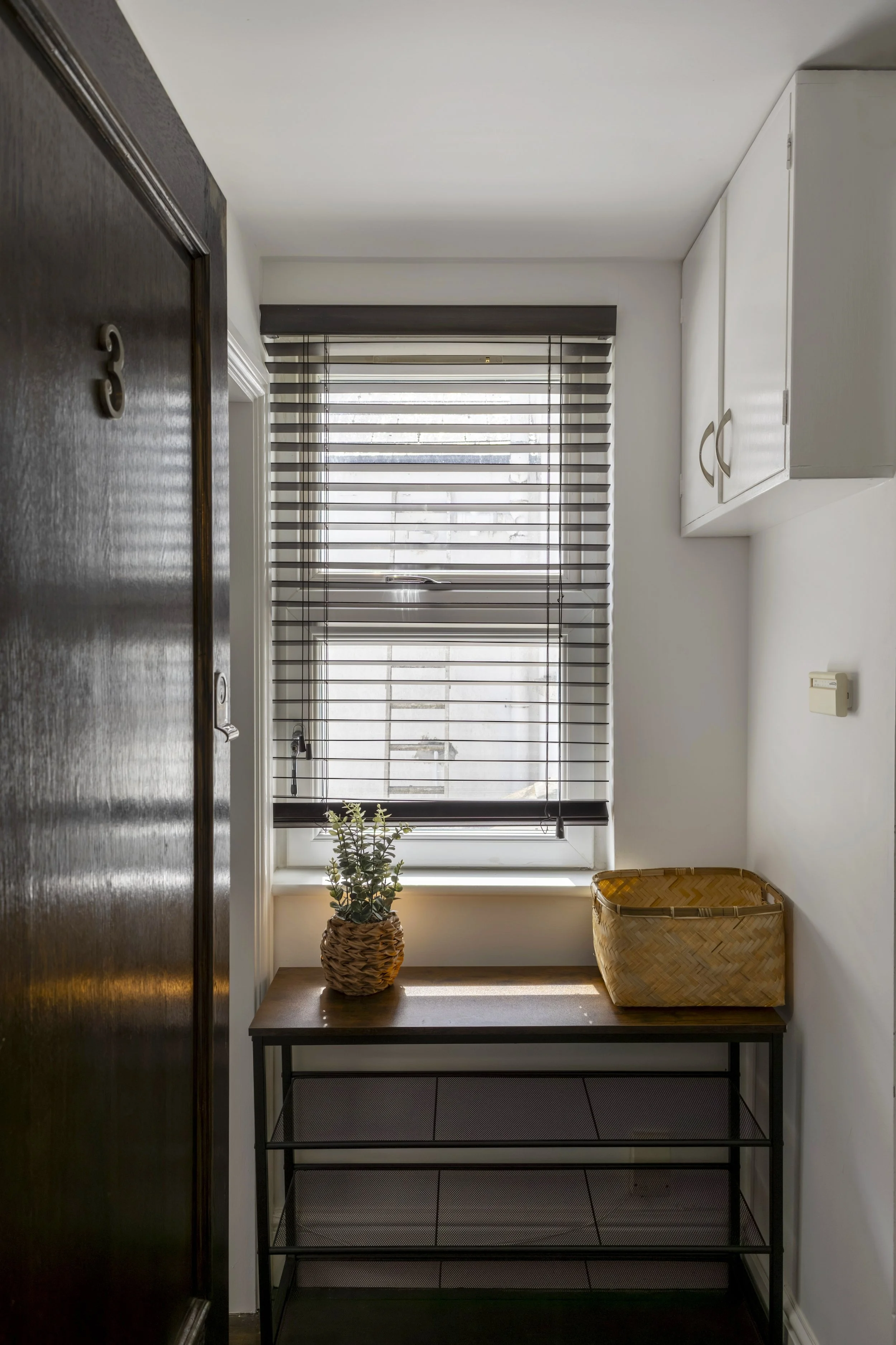 A small table with a potted plant and a empty woven basket in a hallway next to a window with blinds, with a dark door on the left.