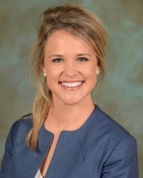 A smiling woman with blonde hair styled in loose waves, wearing a blue blazer over a white top, against a greenish background.