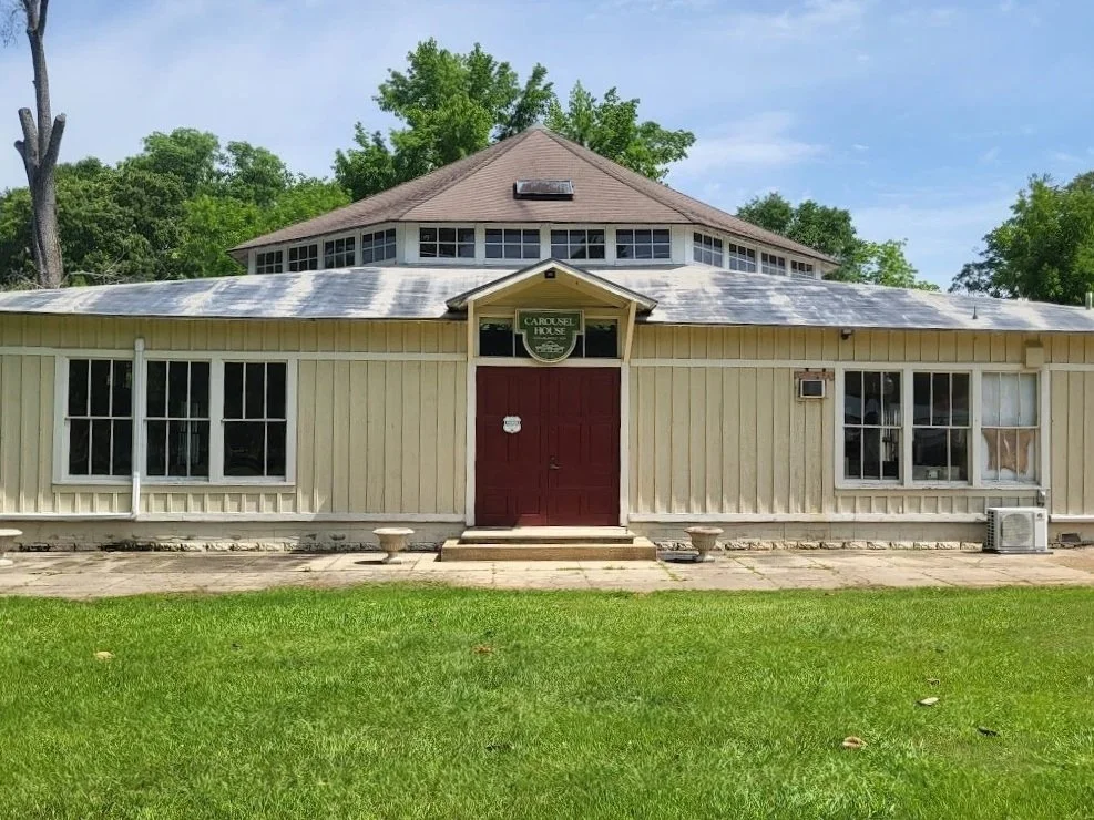 A beige wooden building with a red door, a sign reading 'Carousel House,' and a grassy lawn in front under a blue sky with some clouds.