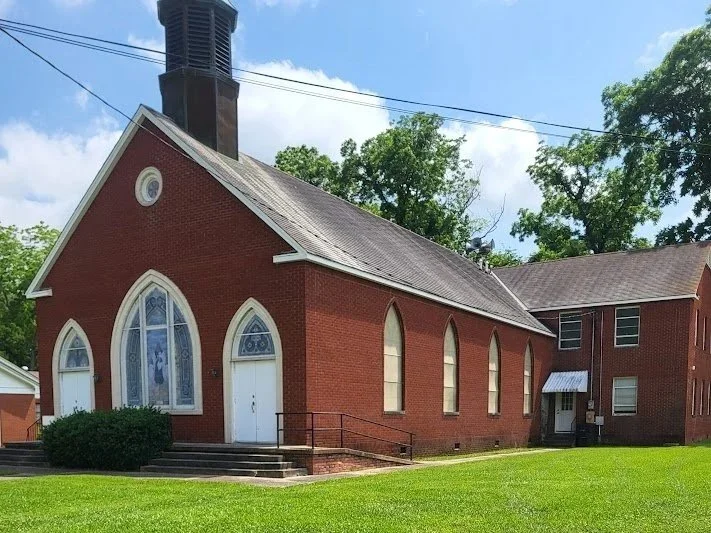 A brick church with pointed arched windows and a tall steeple, surrounded by green grass and trees.