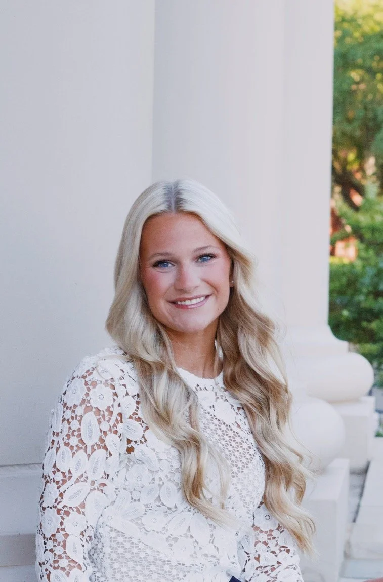A young woman with long, wavy blonde hair, wearing a white lace top, smiling and standing outdoors near white columns with green trees in the background.