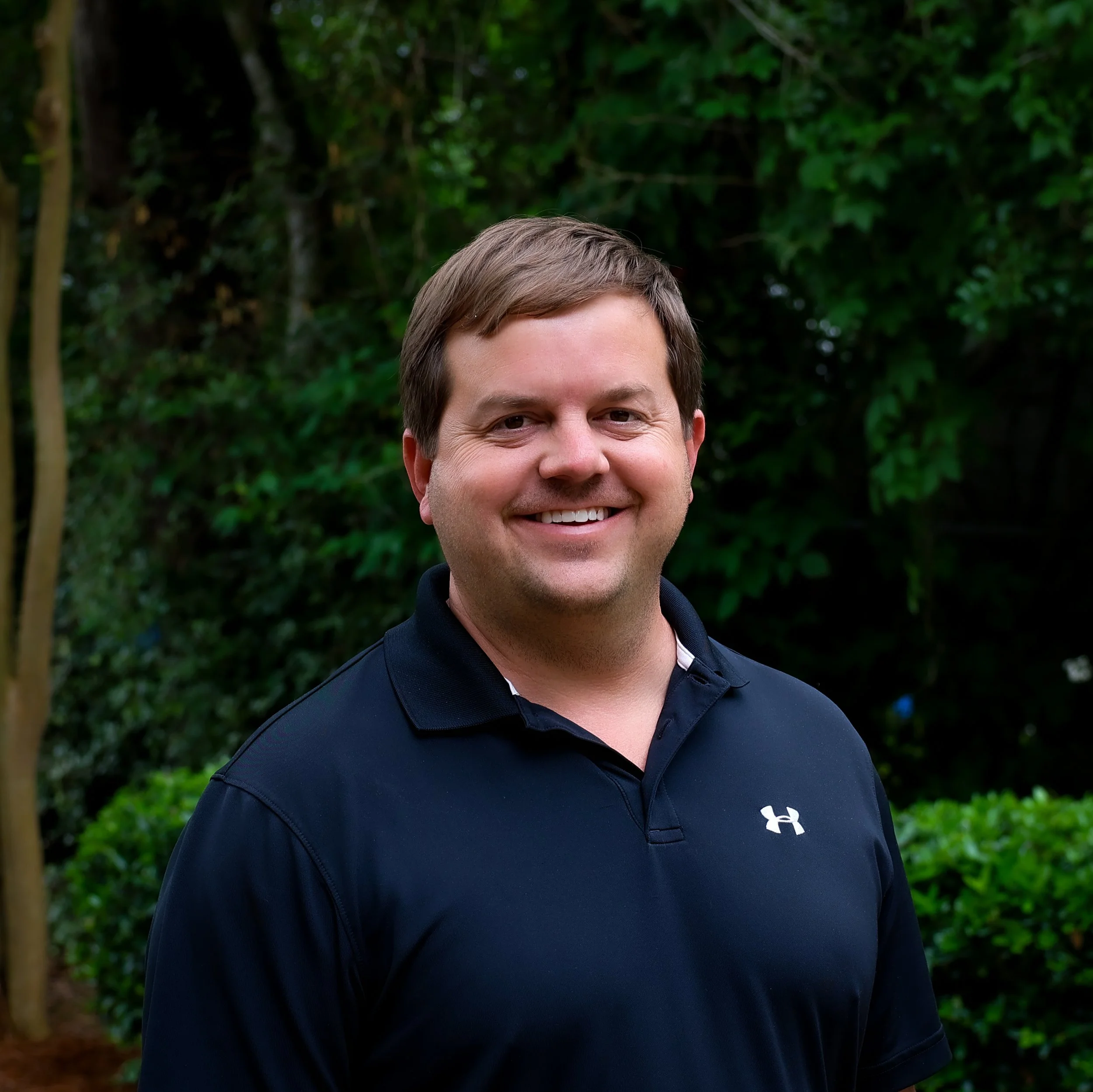 A man with short brown hair and a big smile, wearing a black Under Armour polo shirt, standing outdoors in front of green trees and bushes.