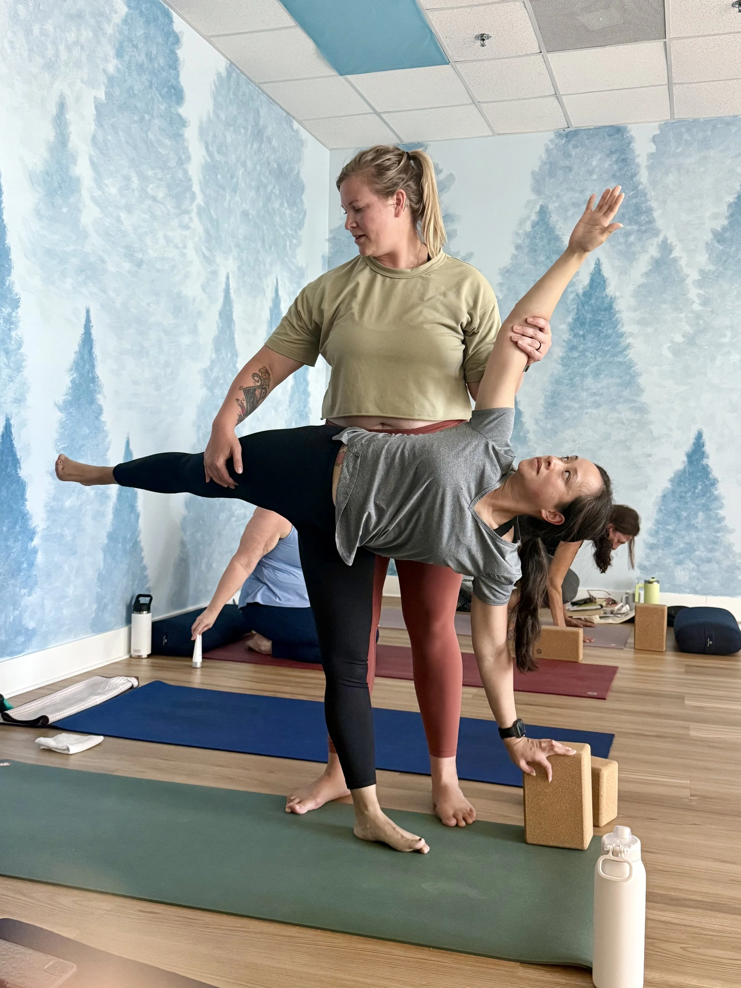 Two women practicing yoga in a studio, with one woman balancing on her left leg while the other supports her by holding her right arm and hip. They are on yoga mats with blocks and water bottles nearby, and others practicing in the background.