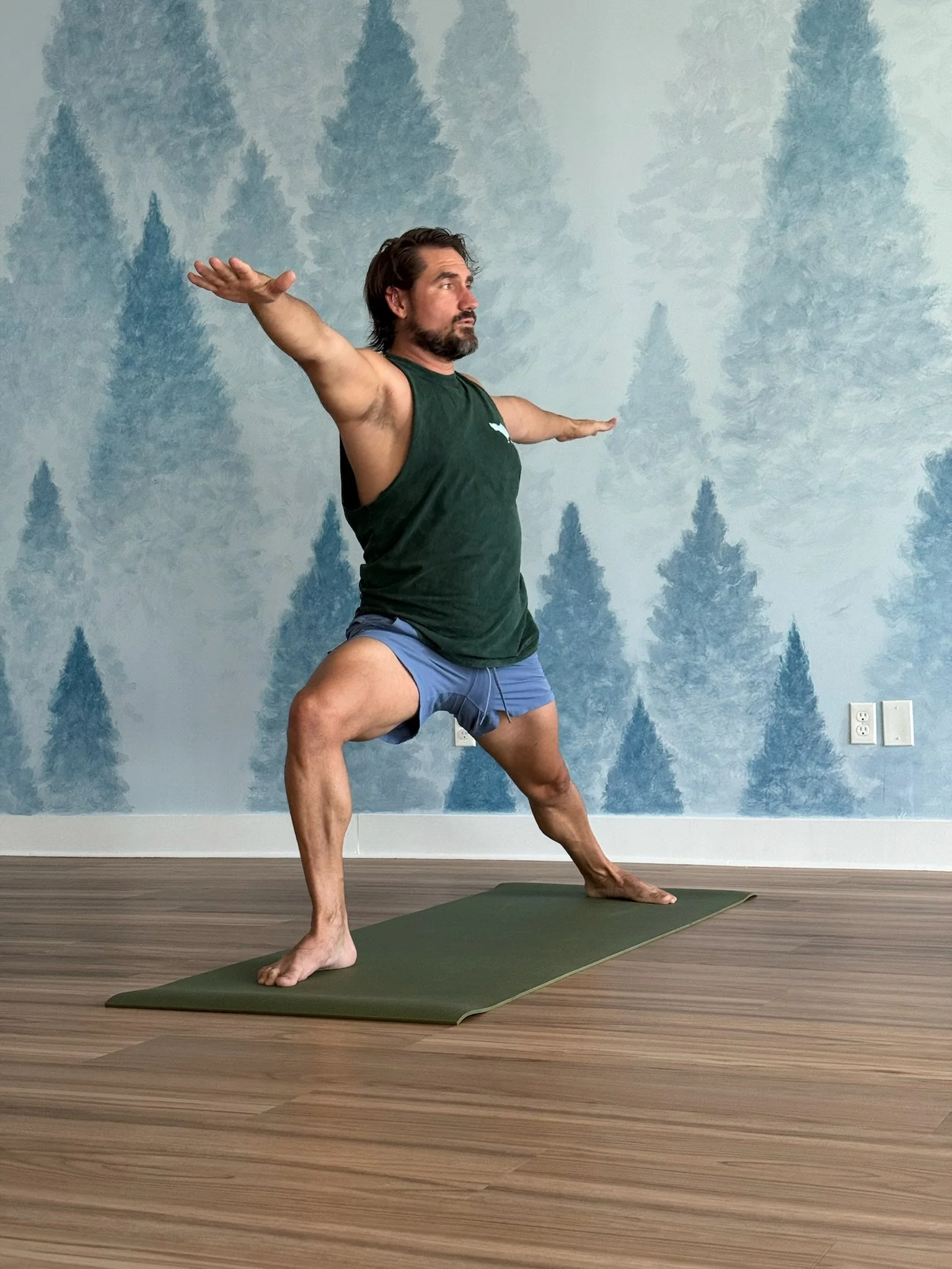 A man practicing yoga indoors, in a lunge pose with one hand on the floor and the other reaching upward, in front of a blue forest-themed wall with potted plants and a small Buddha statue.