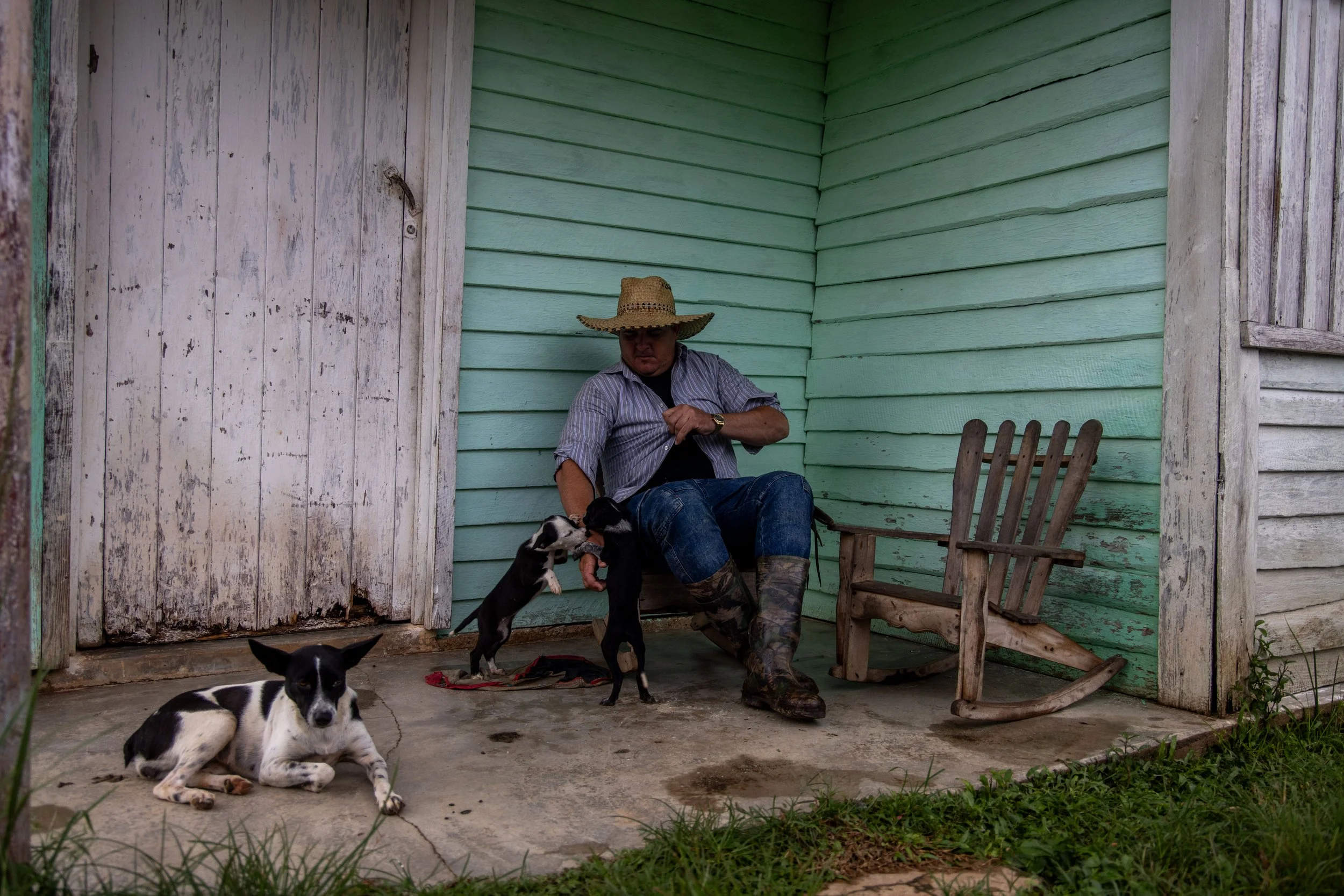 A man wearing a straw hat, striped shirt, and boots, sitting on a wooden chair on a porch, playing with two black and white puppies on a concrete floor outside a weathered house with green and white siding.