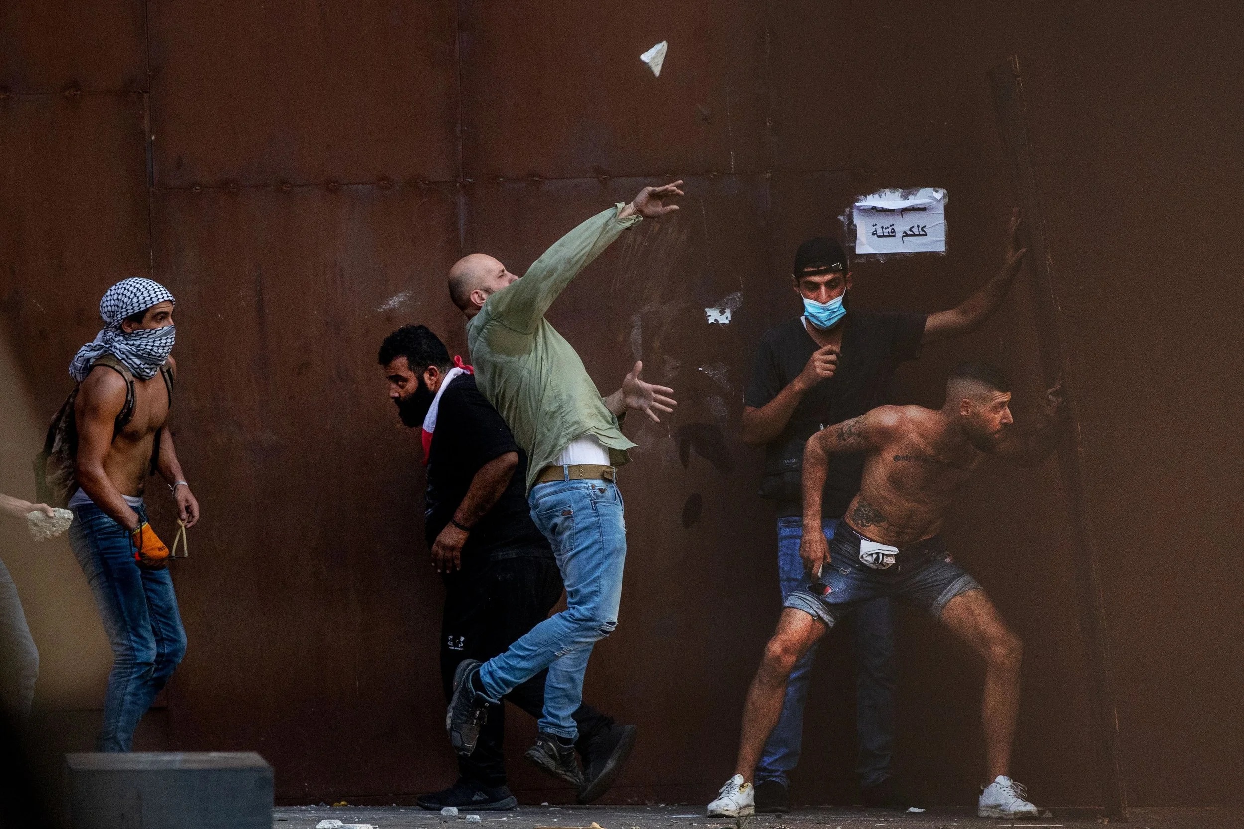 Group of shirtless and masked men protesting, some appear to be fighting or pushing against a metal barrier, one holding a mobile phone, in a tense street scene.