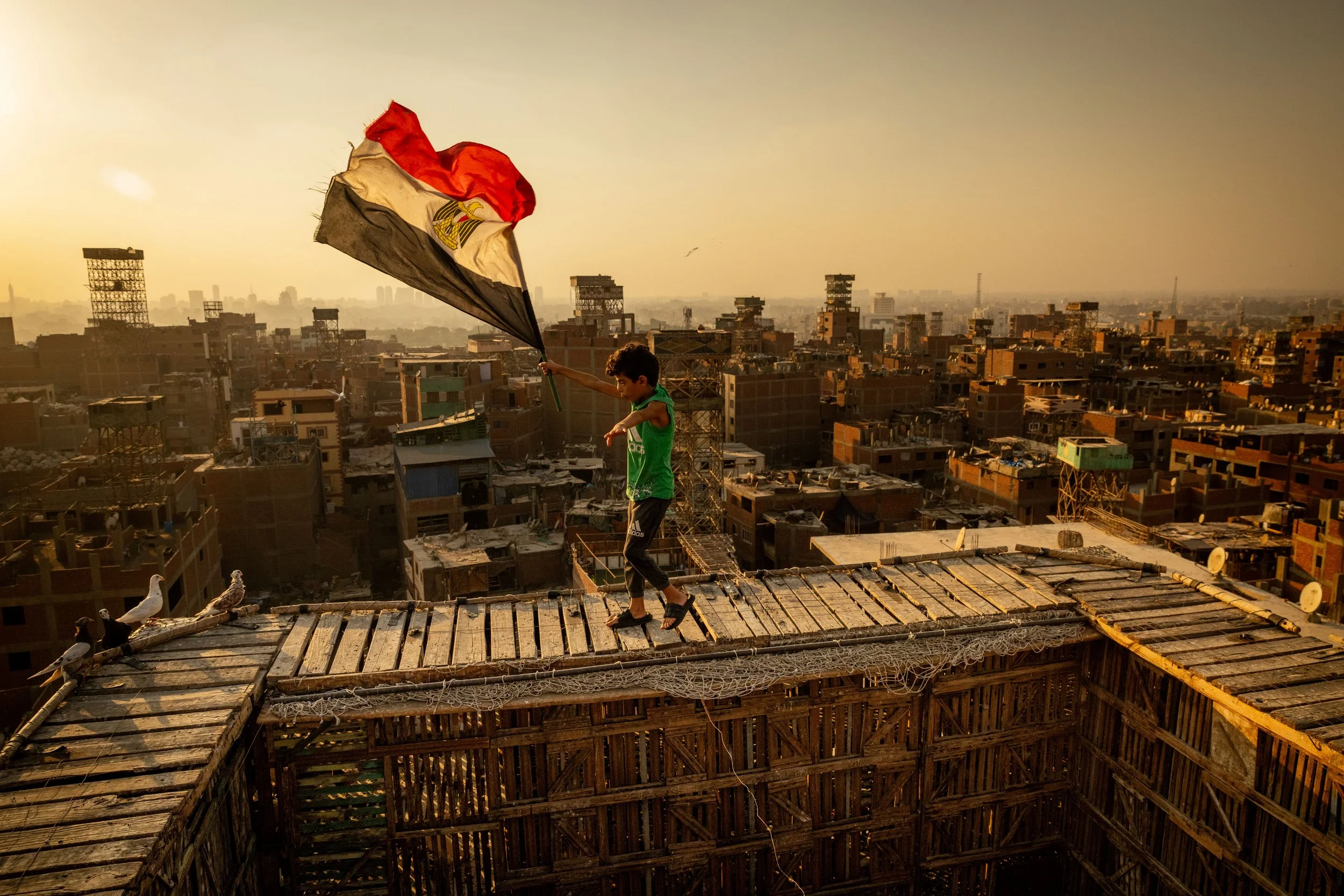 A young boy walking on a makeshift wooden rooftop holding an Egyptian flag with a cityscape in the background during sunset.