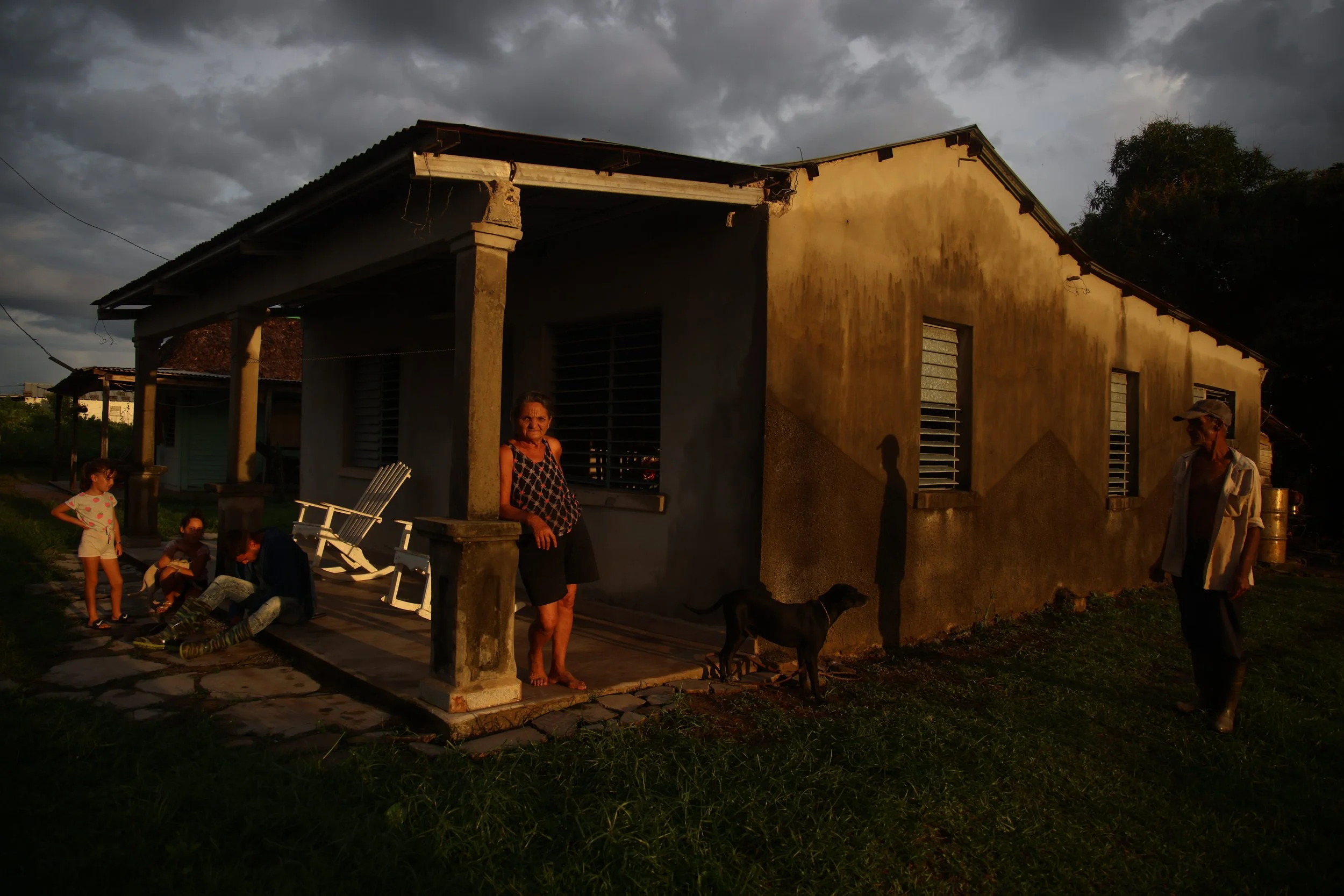 A group of people, including children and adults, standing and sitting outside a yellow house under a cloudy sky during sunset. A black dog is near the house, and shadows are cast on the wall by the sunlight.