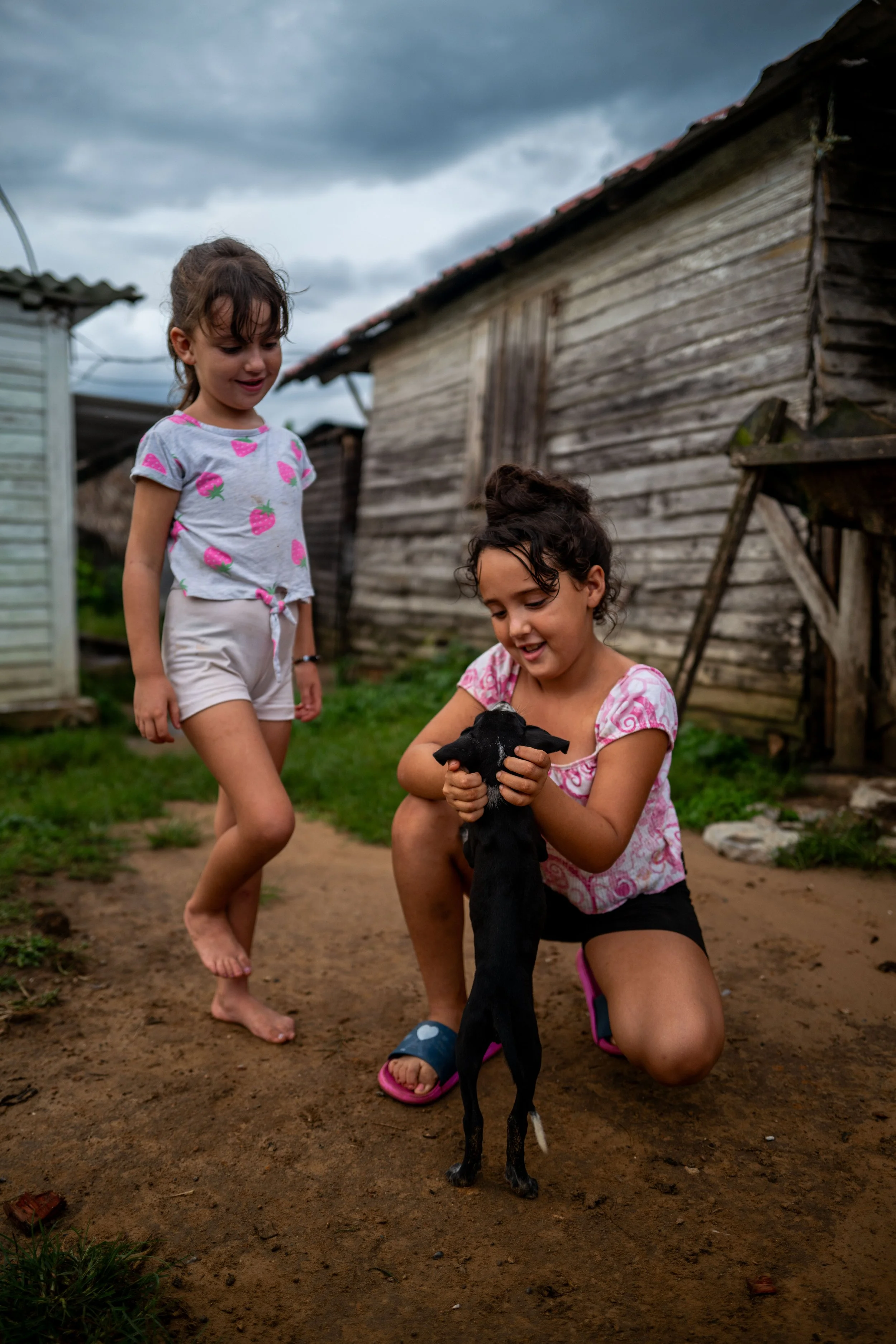 Two young girls playing with a small black puppy outdoors on a dirt path, with old wooden structures and overcast skies in the background.