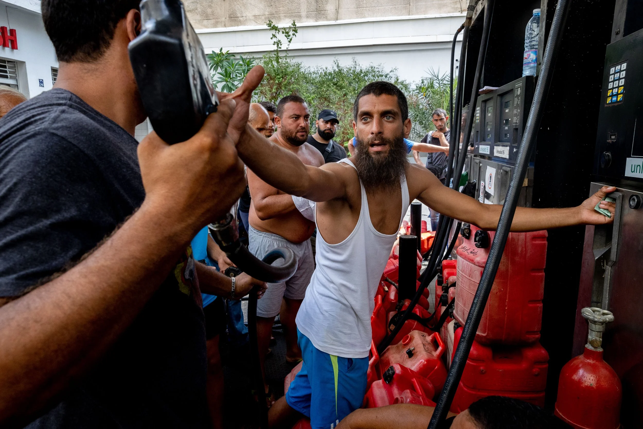 Man with a beard in a white tank top and blue shorts reaching for fuel at a station, surrounded by several people.