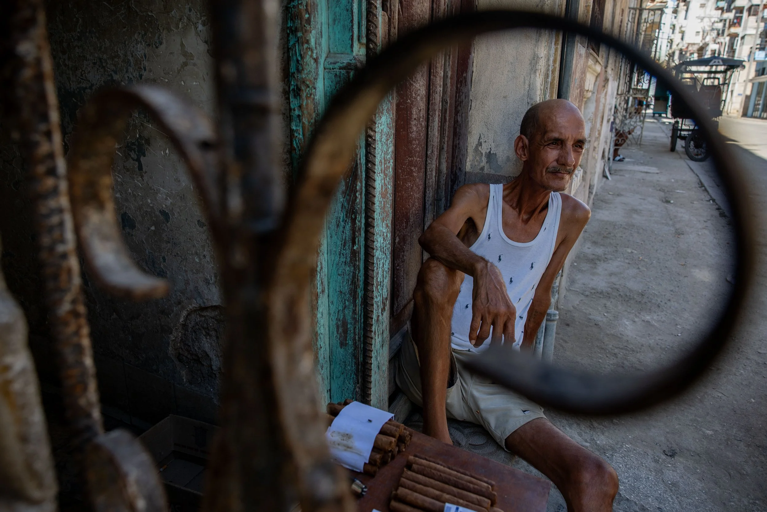 An elderly man with a mustache, wearing a white tank top and beige shorts, sitting on the ground beside a wall on a city street. The photo is taken through a metal fence.