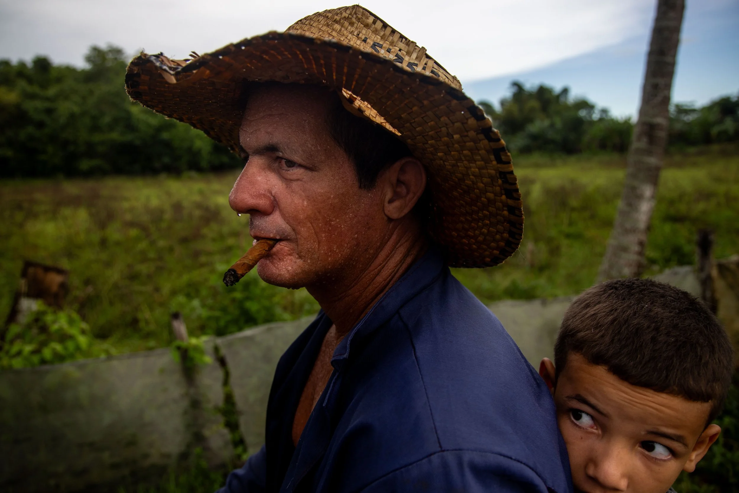 A man with a cigar in his mouth, wearing a straw hat, sitting outdoors with greenery in the background, a young boy riding on his back looking to the side.