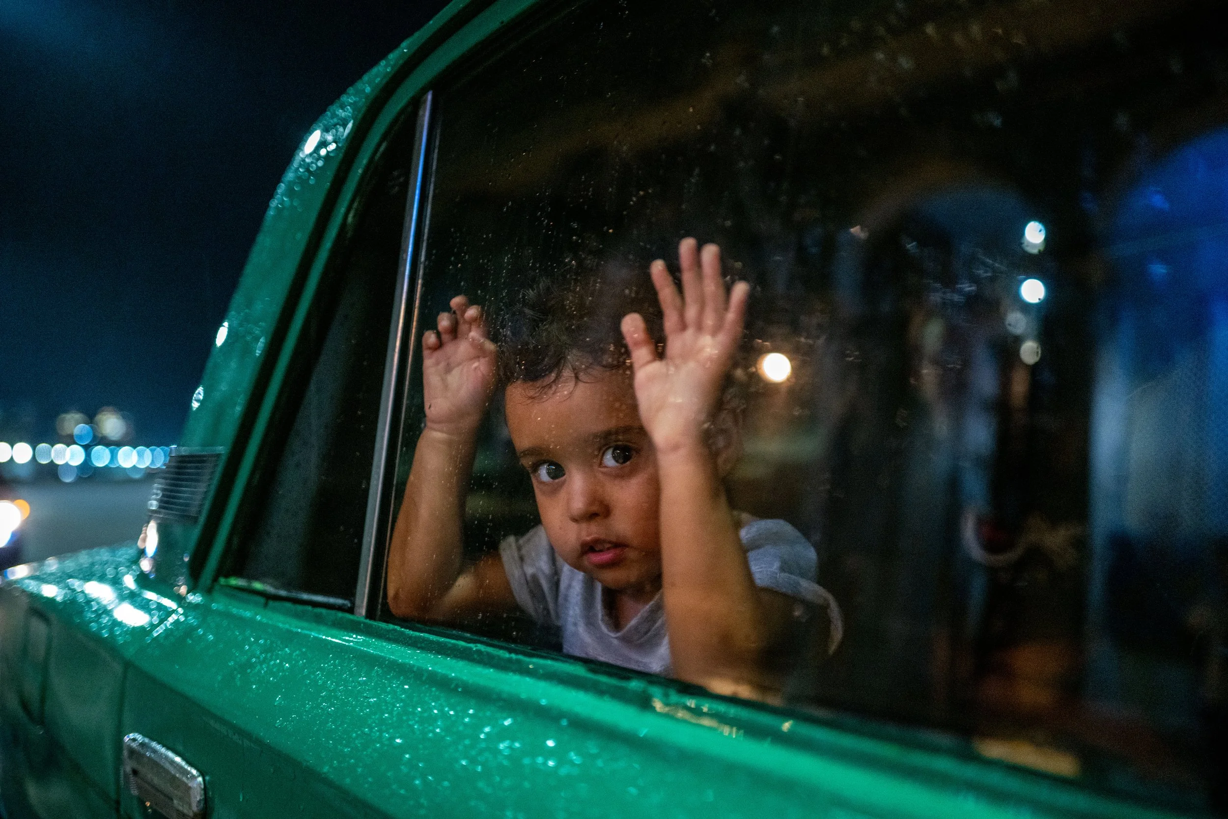 A young child with curly hair looking out of a window of a green vehicle, with hands pressed against the glass, at night.