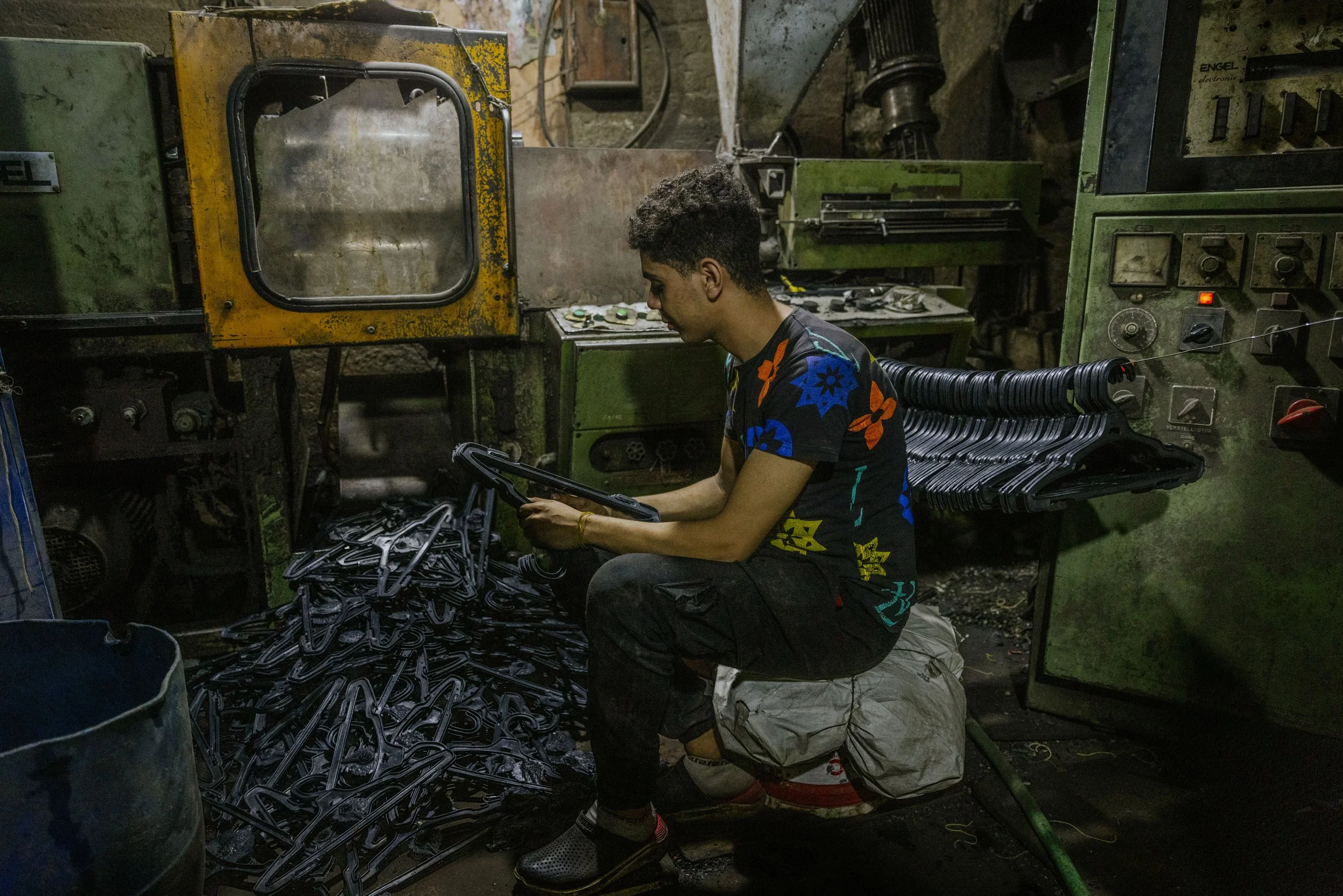 A young man working in a factory with machines and a pile of black plastic hangers.