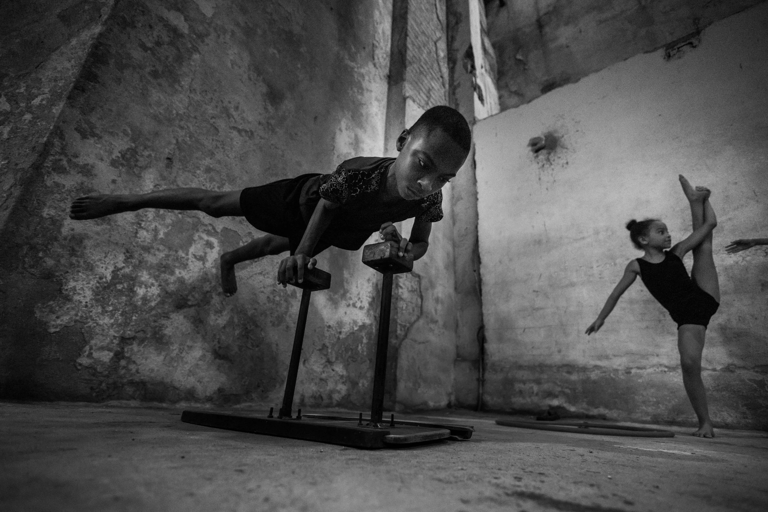 A young boy performing a handstand on parallettes and a young girl practicing a dance move, both in a rustic indoor space with rough concrete walls.