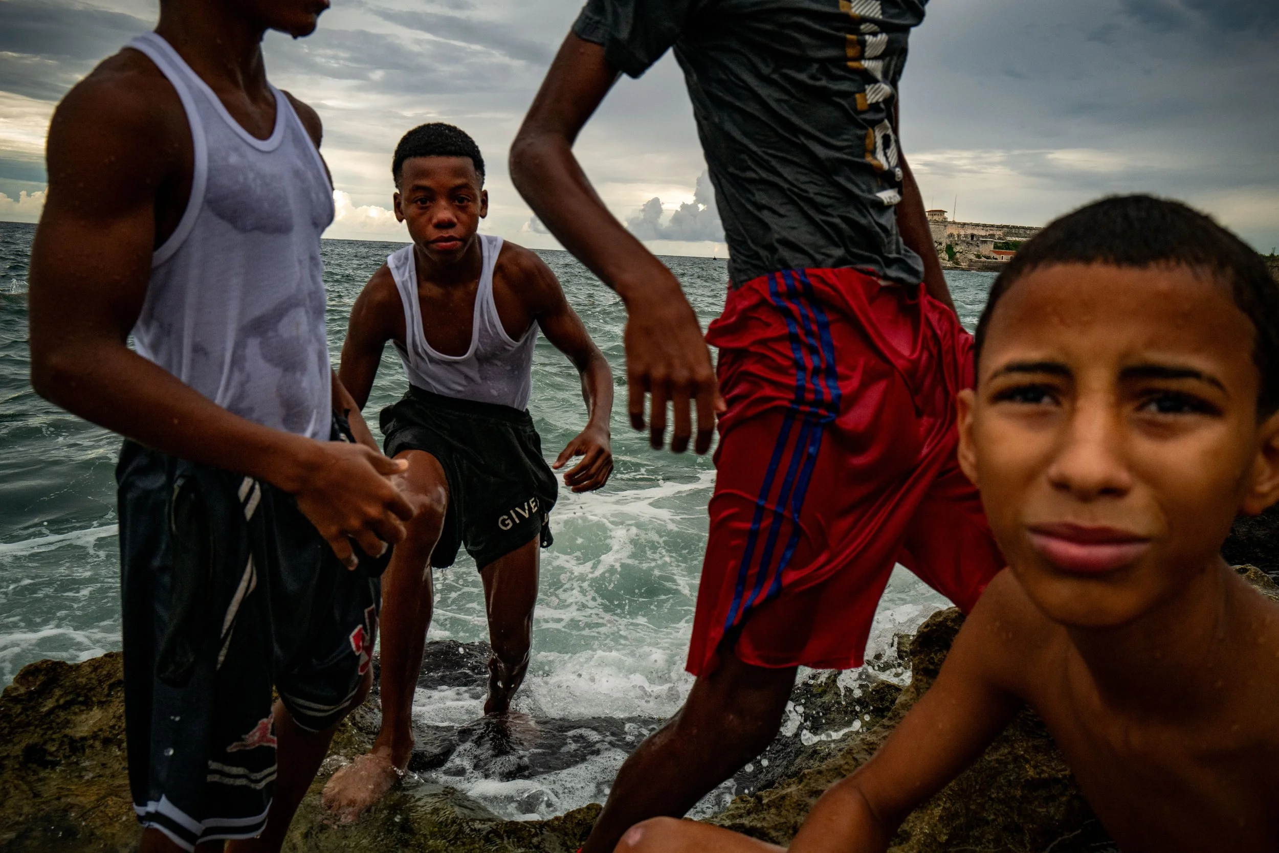 Four boys standing and sitting on rocks by the ocean shoreline on a cloudy day.