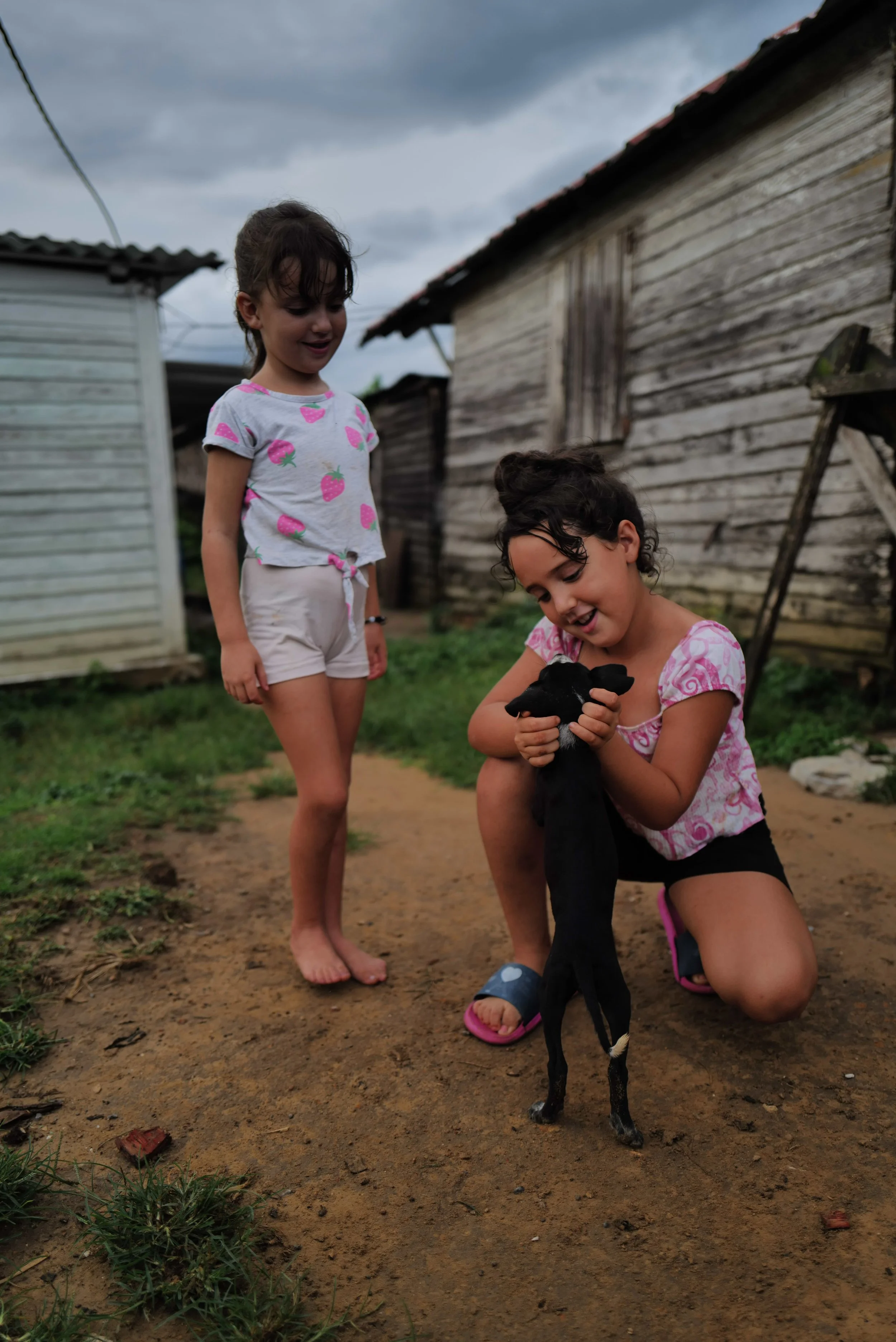 Two young girls playing with a small black goat outside near rustic wooden buildings under a cloudy sky.