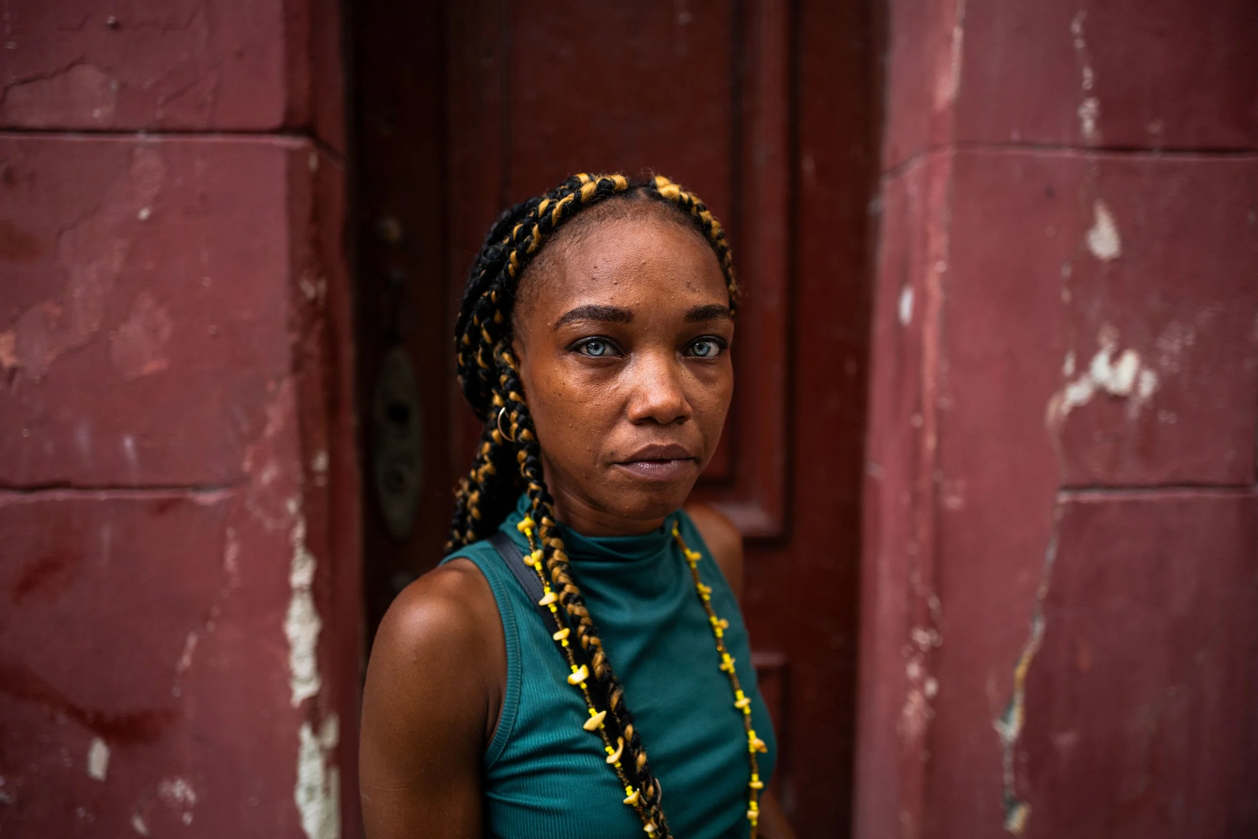 Close-up of a woman with styled braids with yellow beads, gazing at the camera, standing in front of a textured red wall.